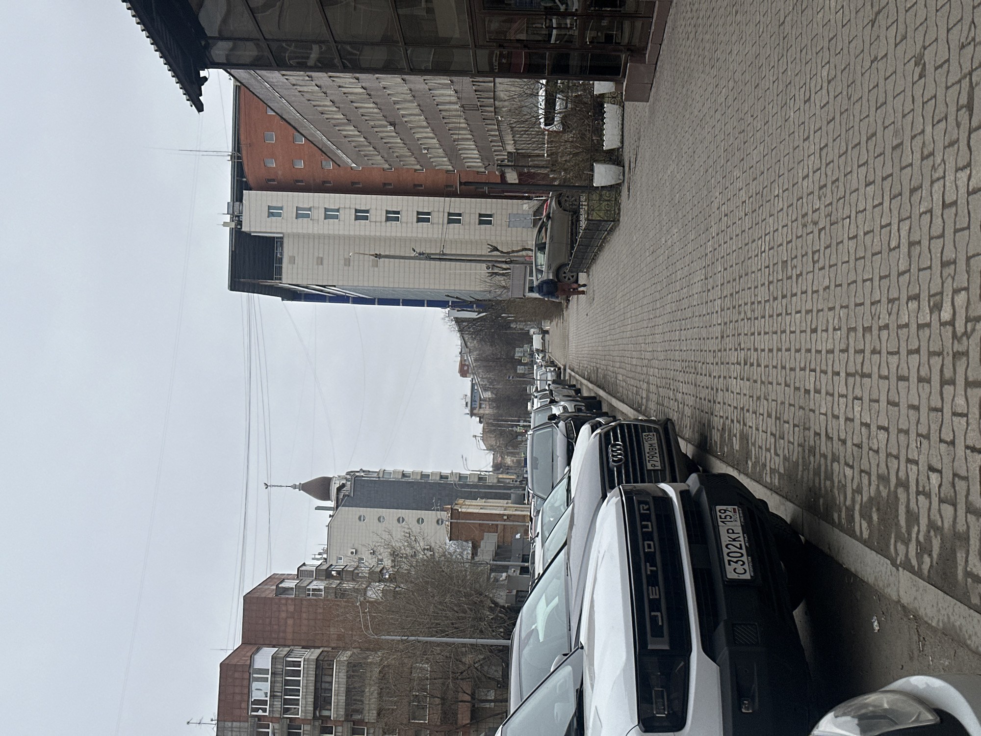 The image shows a street scene in Perm, Russia. Several parked cars line the right side of the street, including a white "JETOUR" SUV with license plate "C302KP 159 RUS". An Audi SUV, with license plate "P790BM 159 RUS", is parked behind it. On the left side of the street, a row of buildings can be seen, with a pedestrian walking alongside a parked car. Overhead cables traverse the street. The street is paved with interlocking concrete pavers. The sky is overcast.