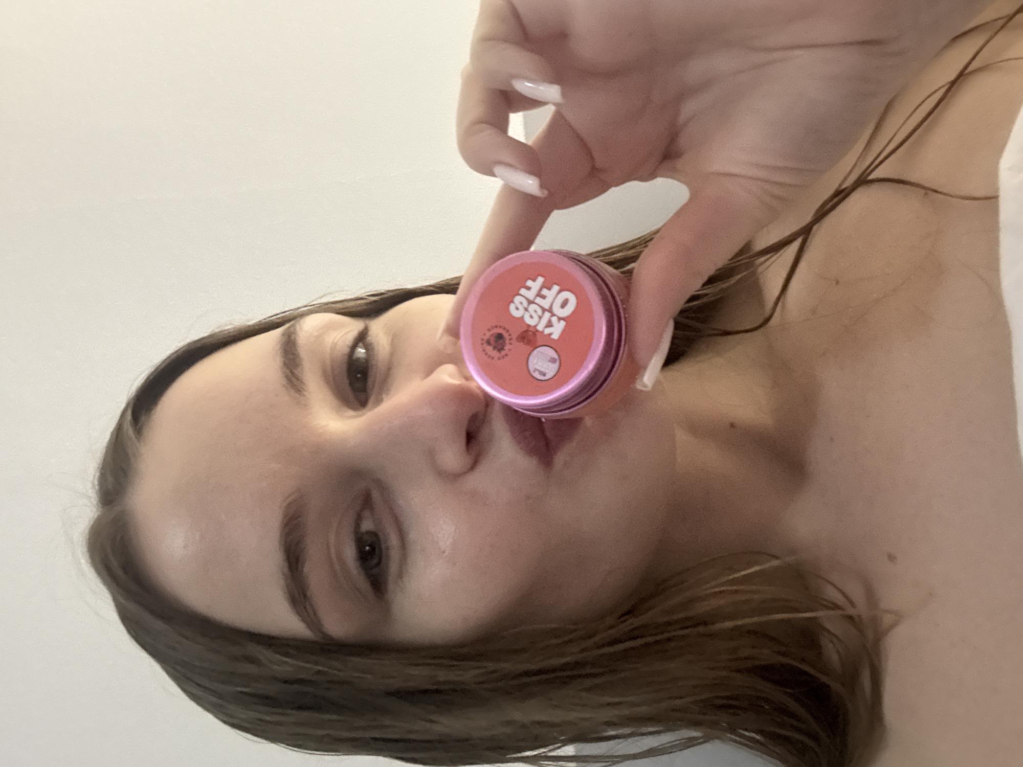A woman is holding a pink container with "KISS OFF" printed on it near her lips. The woman has long, brown hair and pale skin. She has long, manicured fingernails. The background is white and out of focus. There is no indication of this being in Budva, Montenegro.