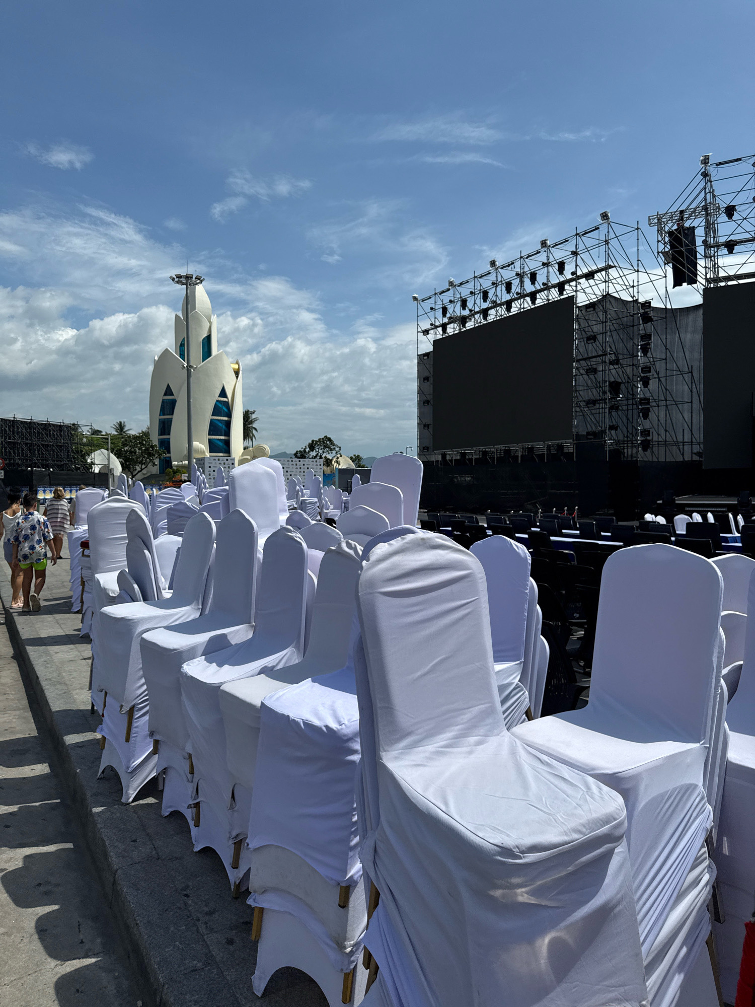 The image depicts an outdoor event setup in Nha Trang, Vietnam, with rows of white-covered chairs set up on a concrete surface. In the background, a large stage structure with a screen and lighting equipment is visible, suggesting an upcoming performance or event. To the left, a distinctive white building with a floral-like design and blue windows stands prominently, characteristic of the Po Nagar Cham Towers complex in Nha Trang. Several people are walking in the background, appearing to be attendees or staff preparing for the event. The sky is blue with scattered clouds, and the scene is illuminated by bright daylight.