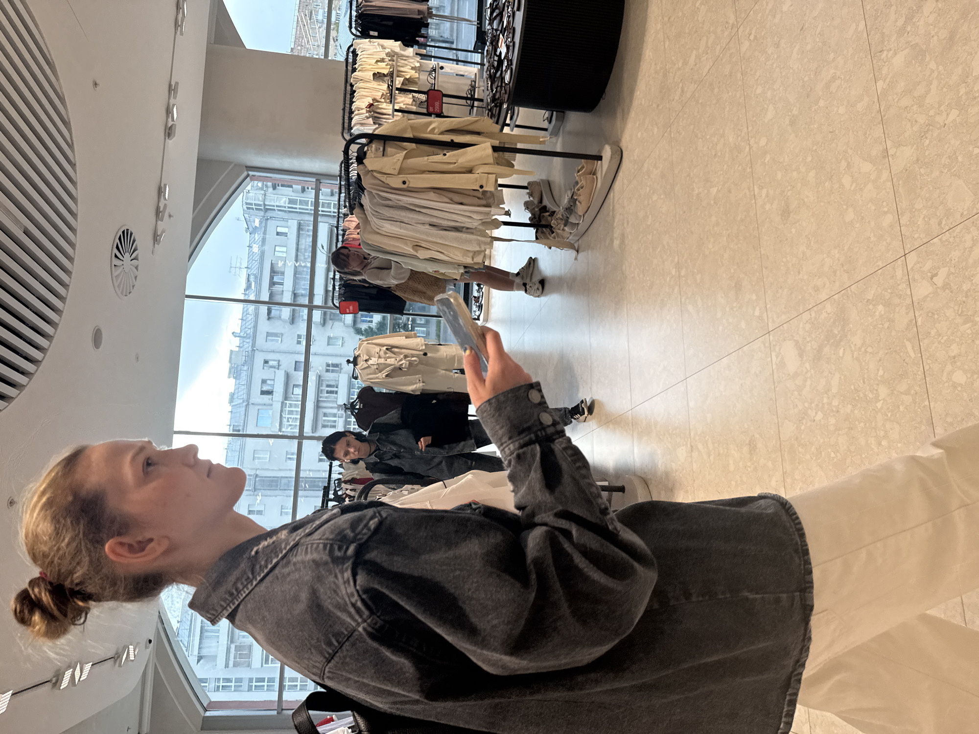 A woman with blonde hair in a bun, wearing a dark denim jacket and white pants, stands in profile in a clothing store, looking at a smartphone in her hand. She is in the foreground, with her left arm extended, holding the phone. In the background, other shoppers are browsing racks of clothing. The store appears modern, with large windows offering a view of urban buildings, likely in Moscow, Russia. The floor is tiled, and clothing racks and displays are visible throughout the space.