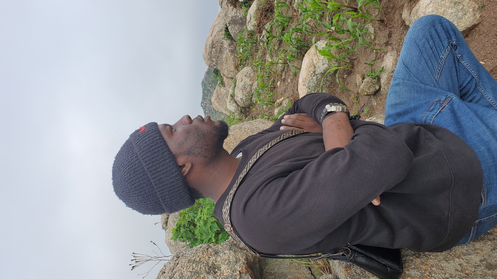 A man wearing a black beanie, black sweatshirt, and blue jeans sits on a rocky outcrop, his arms crossed and his gaze directed to the right. He carries a dark bag slung across his chest. The environment appears to be a natural landscape with large rocks, sparse vegetation, and a slightly overcast sky. The scene is not identifiable as being in Jos, Nigeria.