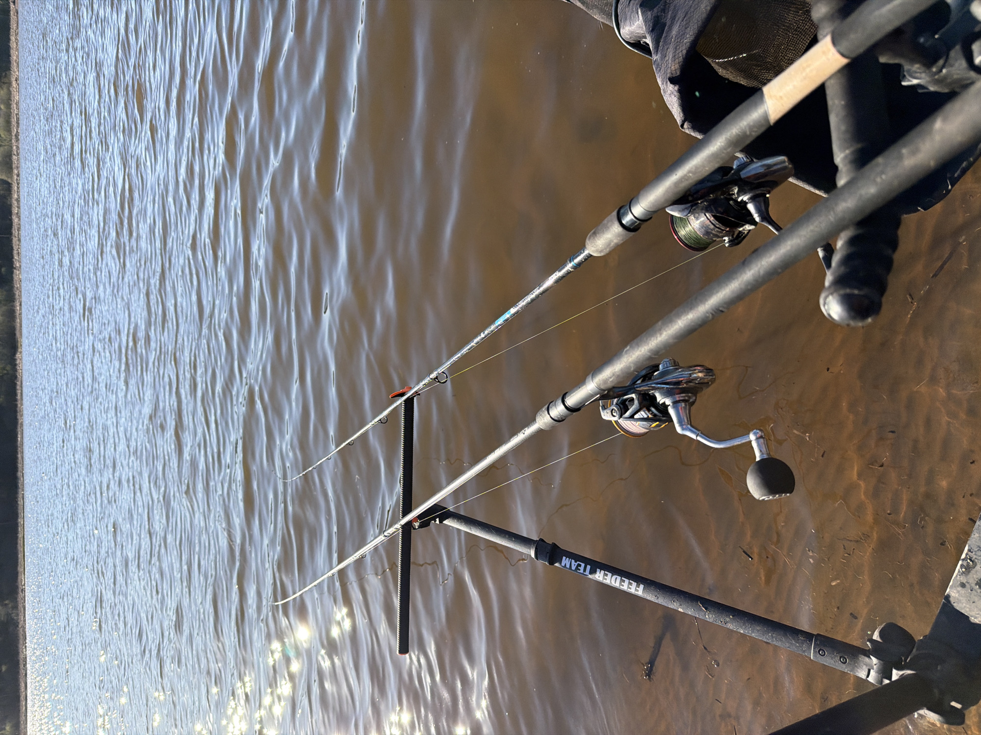 The image depicts two fishing rods set up on a rod rest at a body of water. The rods are equipped with reels, and the fishing line is visible extending into the water. The body of water reflects sunlight and is likely located in Chaykovsky, Russia. The image captures the scene of someone fishing.