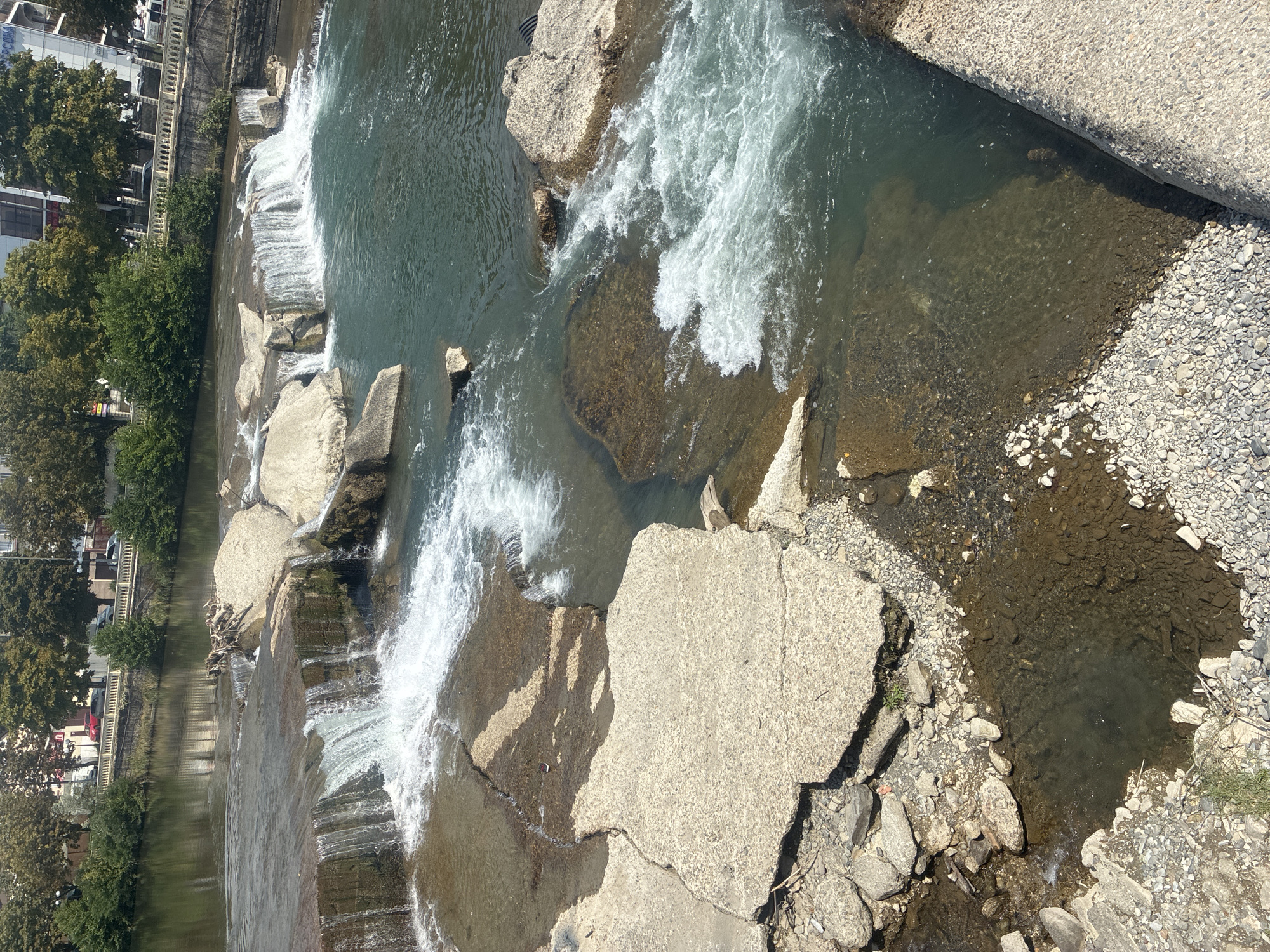 The image depicts a view of the Matsesta River in the city of Sochi, Russia. The river features a series of man-made cascades and shallow waterfalls formed by concrete structures and natural rock formations. The water is clear and appears to be flowing rapidly in some areas, creating white froth. Concrete debris and rounded stones line the riverbed and banks in the foreground. In the background, a tree-lined embankment with a stone retaining wall and a white railing separates the river from a street. A few cars are visible on the street, and buildings and more trees are present further in the background. The sky is bright and appears to be clear.
