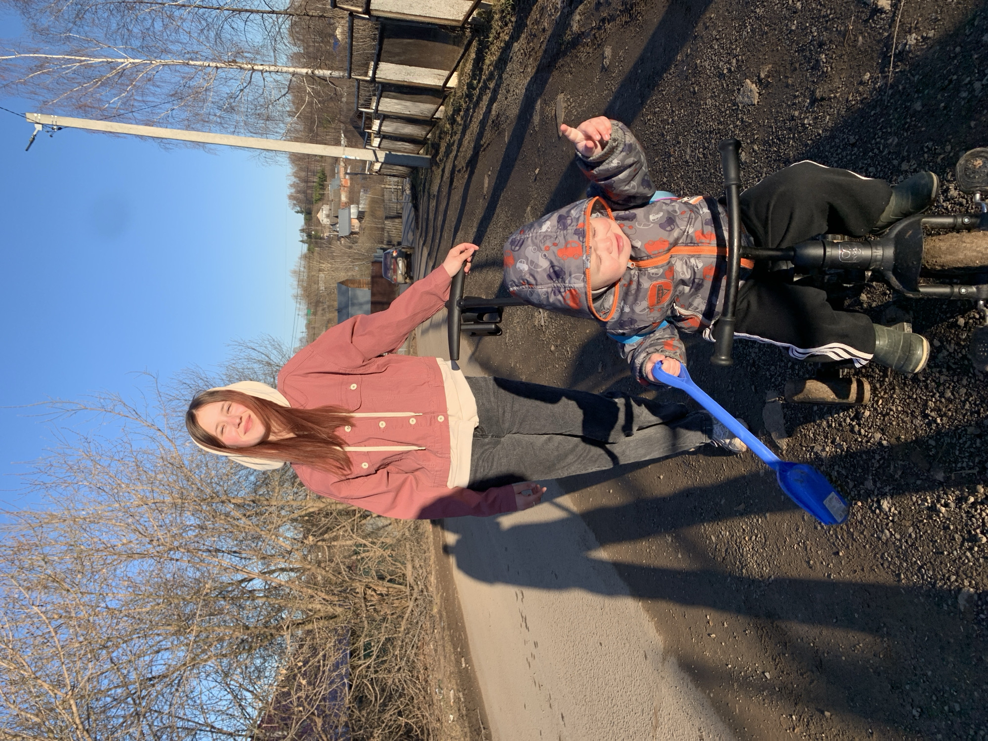 The image depicts two individuals on a tricycle in an outdoor setting. An older adolescent or young adult female in a pink jacket and white hoodie stands behind the tricycle, holding onto its handlebars. A young child, dressed in a patterned winter jacket and black pants with white stripes, sits on the tricycle. A blue plastic shovel is visible near the child's feet.
The ground is a mix of pavement and dirt/gravel. Fences and buildings are visible in the background, as well as trees without leaves. The setting appears to be a street or driveway in a residential area. Based on geolocation, the city is Фоки, Russia.