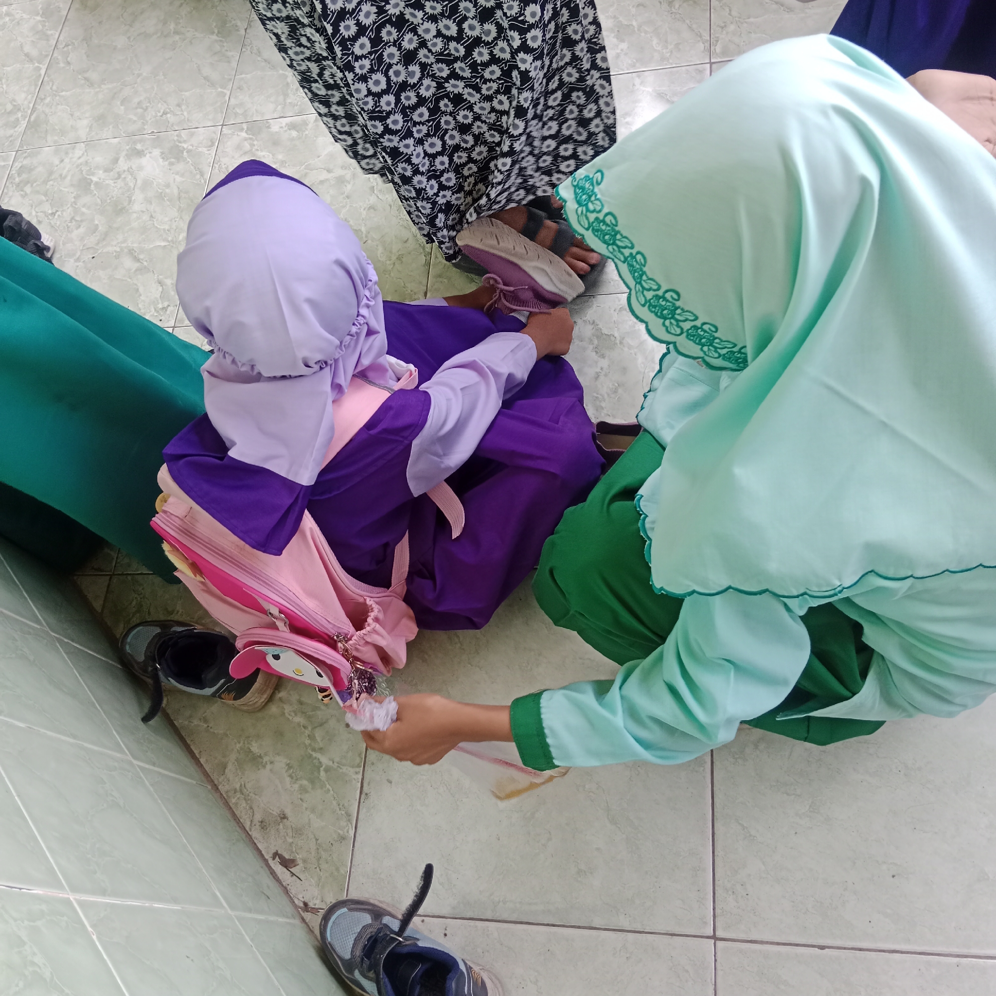 In Tegowanu Kulon, Indonesia, three individuals, two children and one adult, are positioned on a tiled floor. A young child, wearing a light purple hijab and a purple and pink outfit with a backpack, is seated. To their right, a woman or older child, in a mint green hijab and green top, is leaning down and handing a white tissue or napkin to the younger child, who is holding a clear plastic bag. Another person, partially visible in a black and white floral dress and grey sandals, stands to the upper left. The environment appears to be an outdoor or indoor space with light-colored tiles. A green garment is visible to the left, and several pairs of shoes are scattered around.