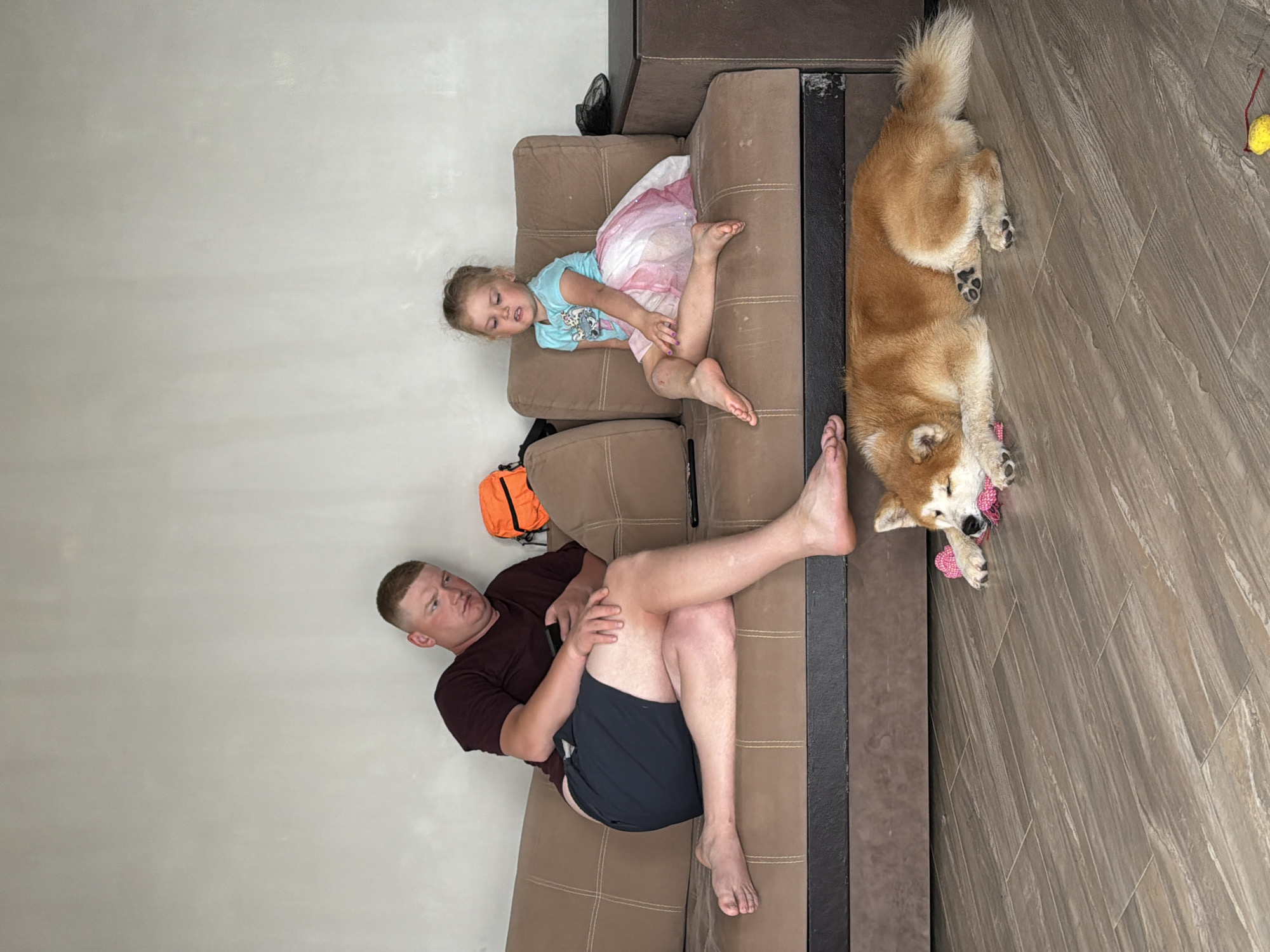 A photograph shows a man, a girl, and a dog in a living room in Chaikovsky, Russia. The man is reclining on a brown couch, wearing a maroon shirt and navy shorts. A small girl is lying on the couch to the left of the man, wearing a blue shirt and a pink and white tutu. An orange backpack rests on the couch near the girl. A dog, tan and white in color, lies on the floor near the edge of the couch, with a pink toy in its paws. The floor has a gray wood grain pattern. A yellow ball is near the dog, and a black object is visible on the back of the couch. The walls are light gray.