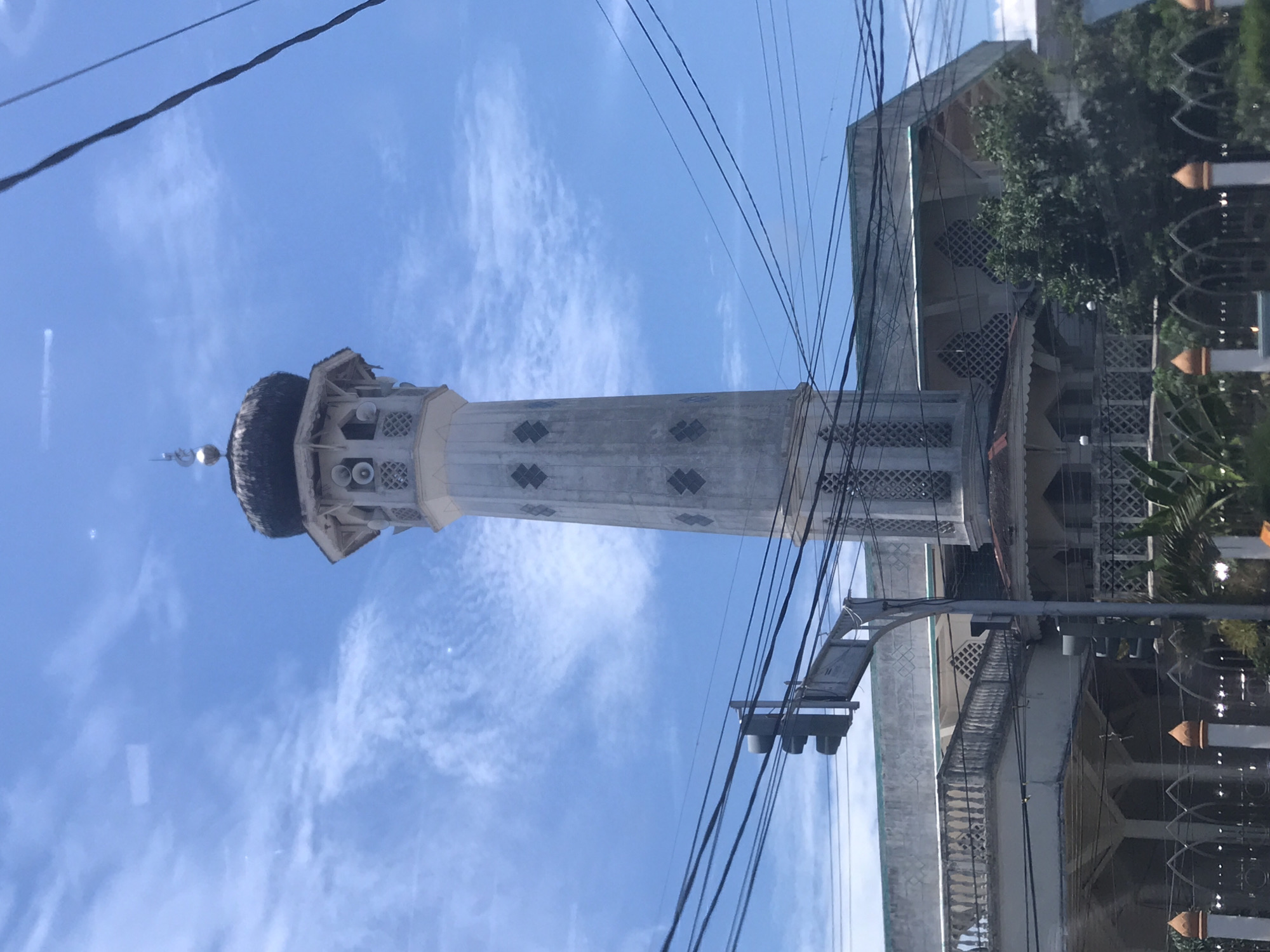 The image shows a minaret of a mosque in Lhokseumawe, Indonesia, against a blue sky with light clouds. Power lines intersect the view of the minaret, which is tall and cylindrical with a decorated top. Part of the mosque building is visible on the right, featuring detailed architectural patterns. A street light and foliage are also present on the right side of the frame.