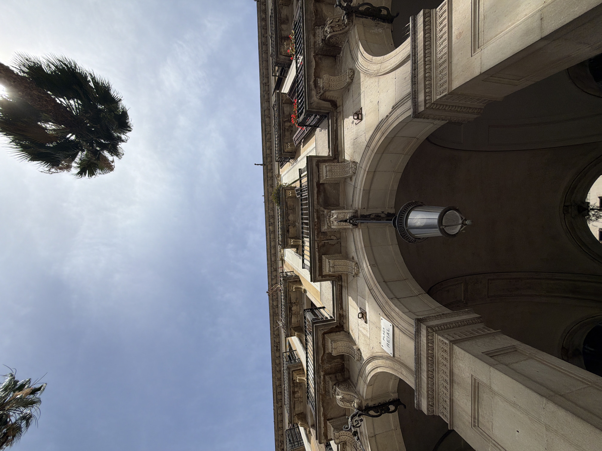An upward-angle shot captures the ornate architecture of the Plaça Reial in Barcelona, Spain, on a bright, partly cloudy day. The building facade features multiple levels of balconies with decorative iron railings, supported by elaborate stone corbels. Archways form a covered walkway at street level, with a hanging lamp visible beneath one of them. A sign on the wall reads "PLAÇA REIAL." Palm trees are visible at the top of the frame, their fronds silhouetted against the sky. The sun is bright, creating some lens flare. No people or animals are present in the image.