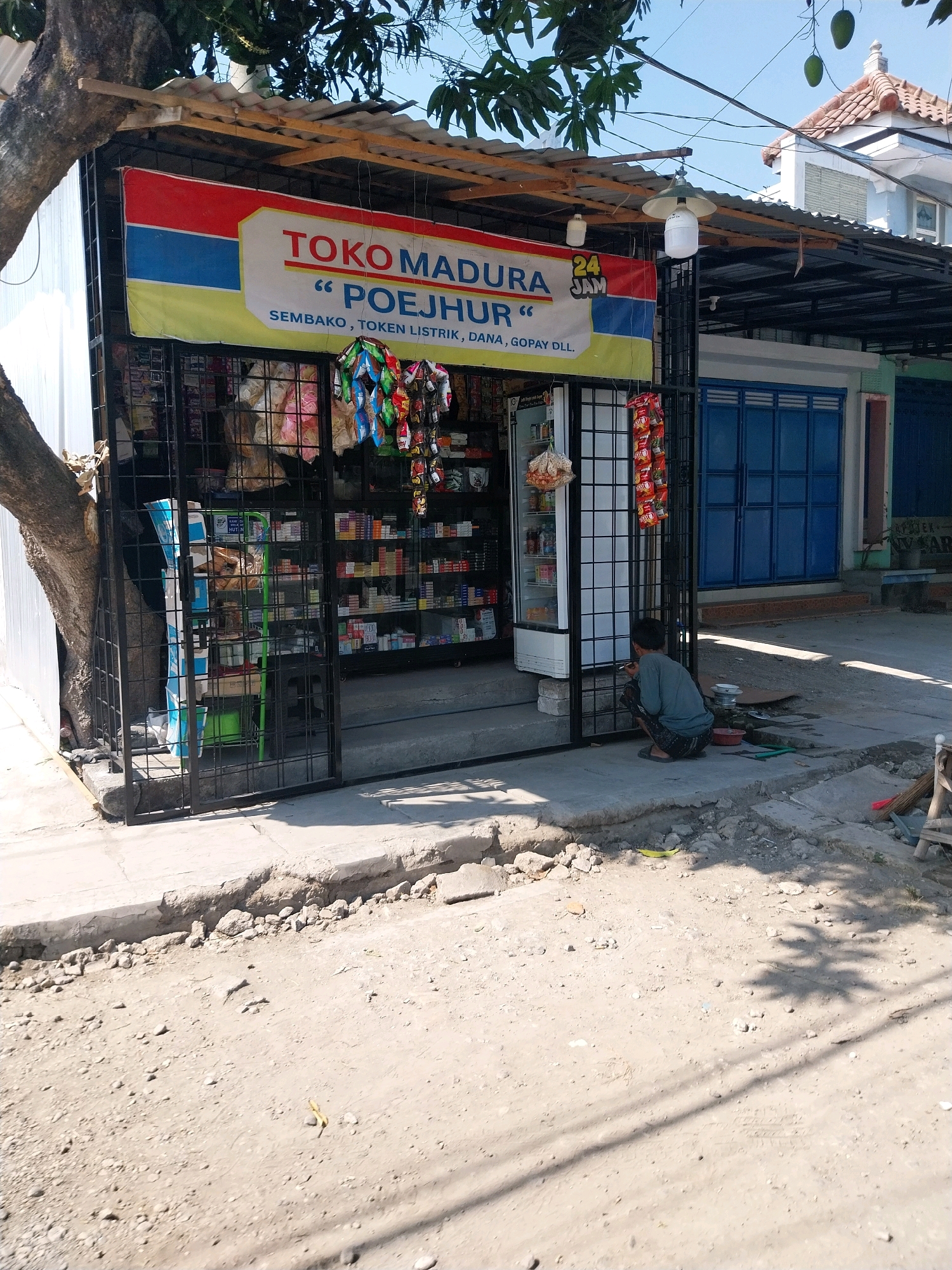 The image depicts a small shop, identified by a banner as "TOKO MADURA POEJHUR", located in Pandean, Indonesia. The shop appears to be a convenience store, with various goods displayed behind a metal gate. Products visible include packaged snacks, drinks, and what appear to be cigarettes. The shop has a corrugated metal roof and is attached to a building with blue doors. A person is crouched on the sidewalk in front of the shop, seemingly engaged in some activity. The surrounding environment appears to be a dusty street or pathway with some rubble. The lighting suggests it is daytime, with shadows cast by trees and overhead structures.