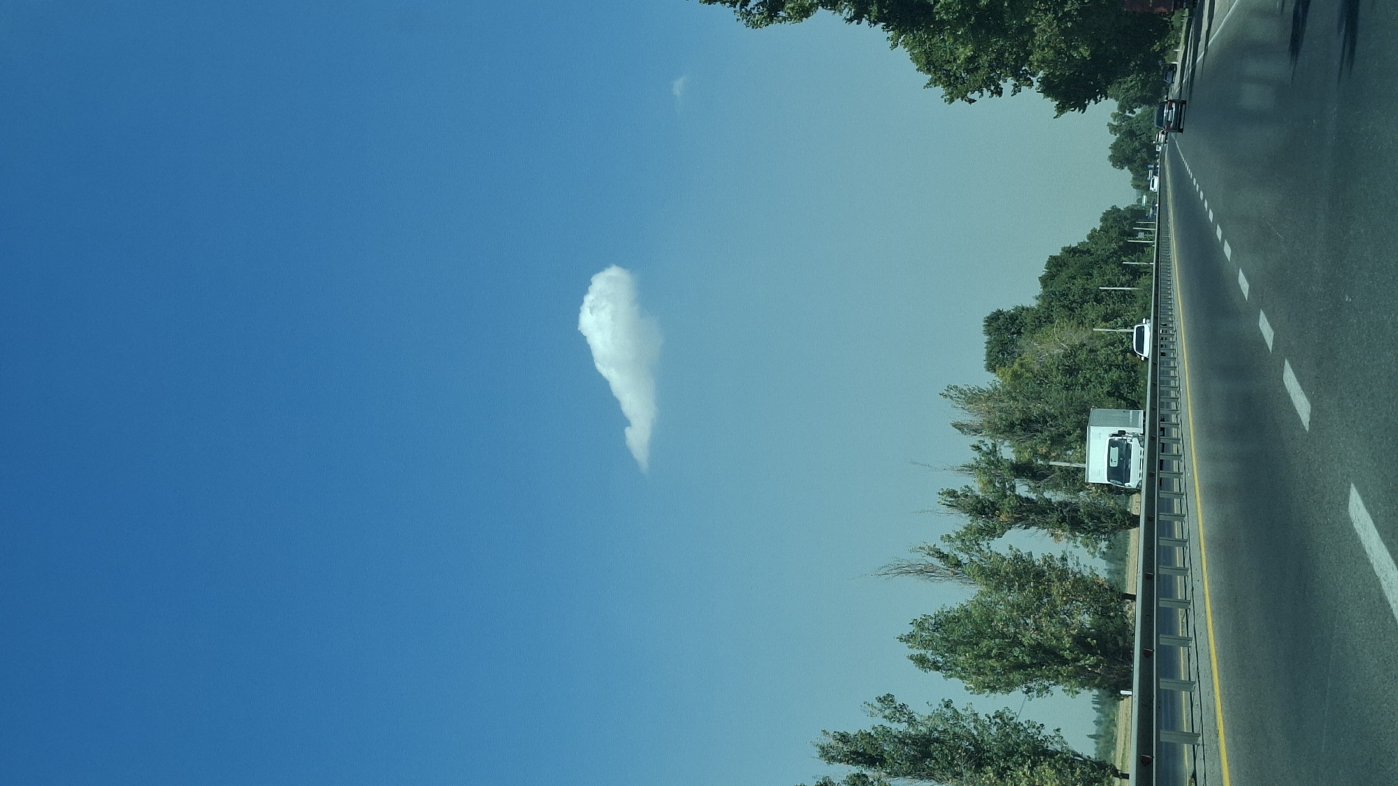 The image depicts a highway scene with a clear blue sky. A single, elongated white cloud is visible in the upper portion of the sky. The highway stretches into the distance, with lane markings clearly visible. A metal guardrail separates the highway from a grassy area with trees. A white box truck is seen on the highway, along with other vehicles further down the road. The trees on the side of the road are lush and green, suggesting a warm season. The overall environment appears to be a rural or suburban area, with no specific landmarks indicating a precise city location.