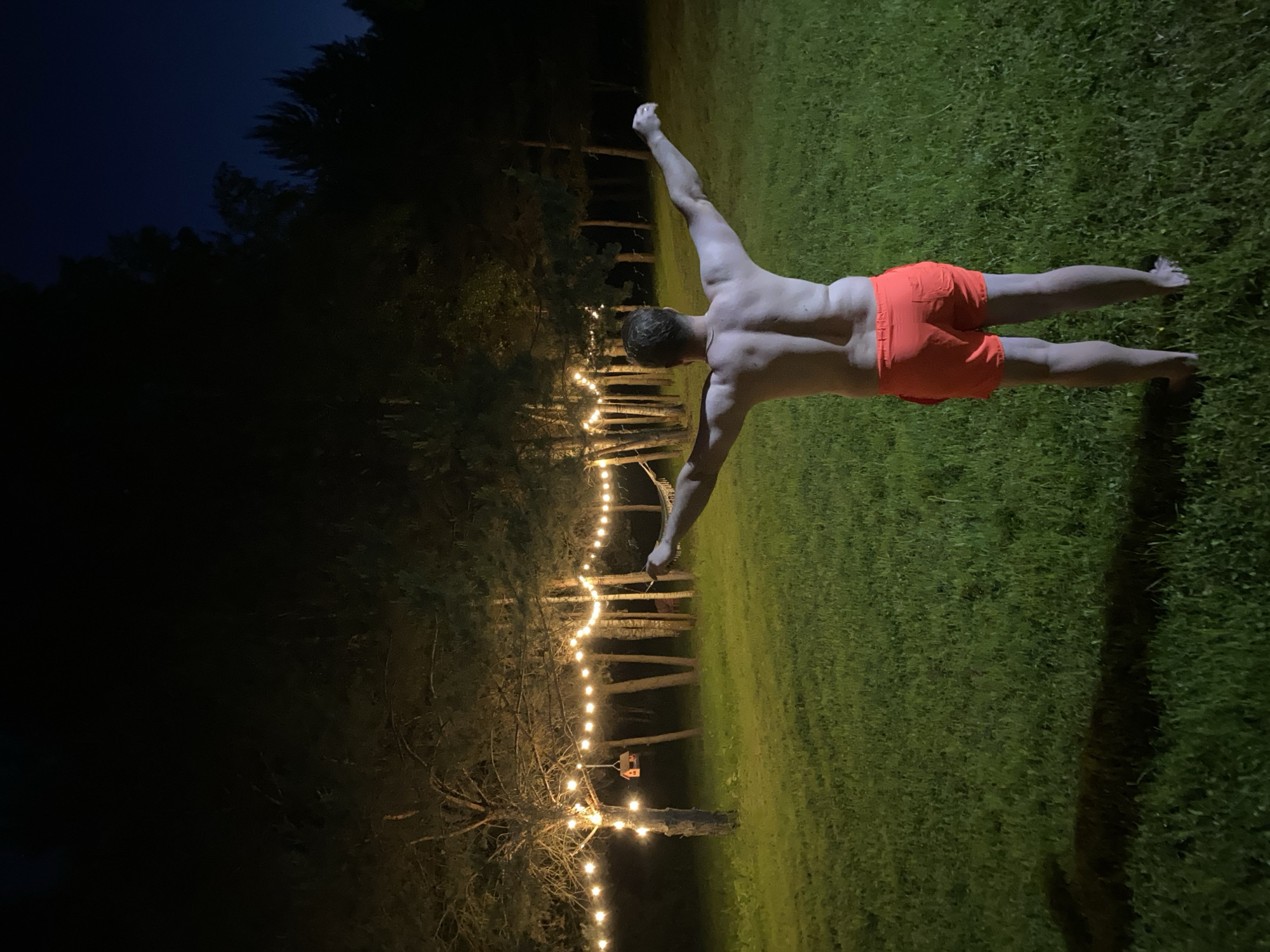 The image depicts a nighttime outdoor scene in what is stated to be Копнино, Russia. A lone adult male, viewed from behind, stands barefoot on a manicured grass lawn. He is shirtless, exposing his back, and wears bright orange shorts. His arms are extended horizontally outwards from his sides. In the background, numerous trees, predominantly pines and some possibly birches, form a sparse forest line. A string of warm-toned incandescent light bulbs is strung horizontally between several trees, illuminating the immediate area. A small, orange-roofed birdhouse is suspended from a tree trunk on the left side of the frame. To the right, partially visible behind the man's outstretched right arm, is a white or light-colored hammock hanging between two trees. The sky above is dark, indicating a clear night.