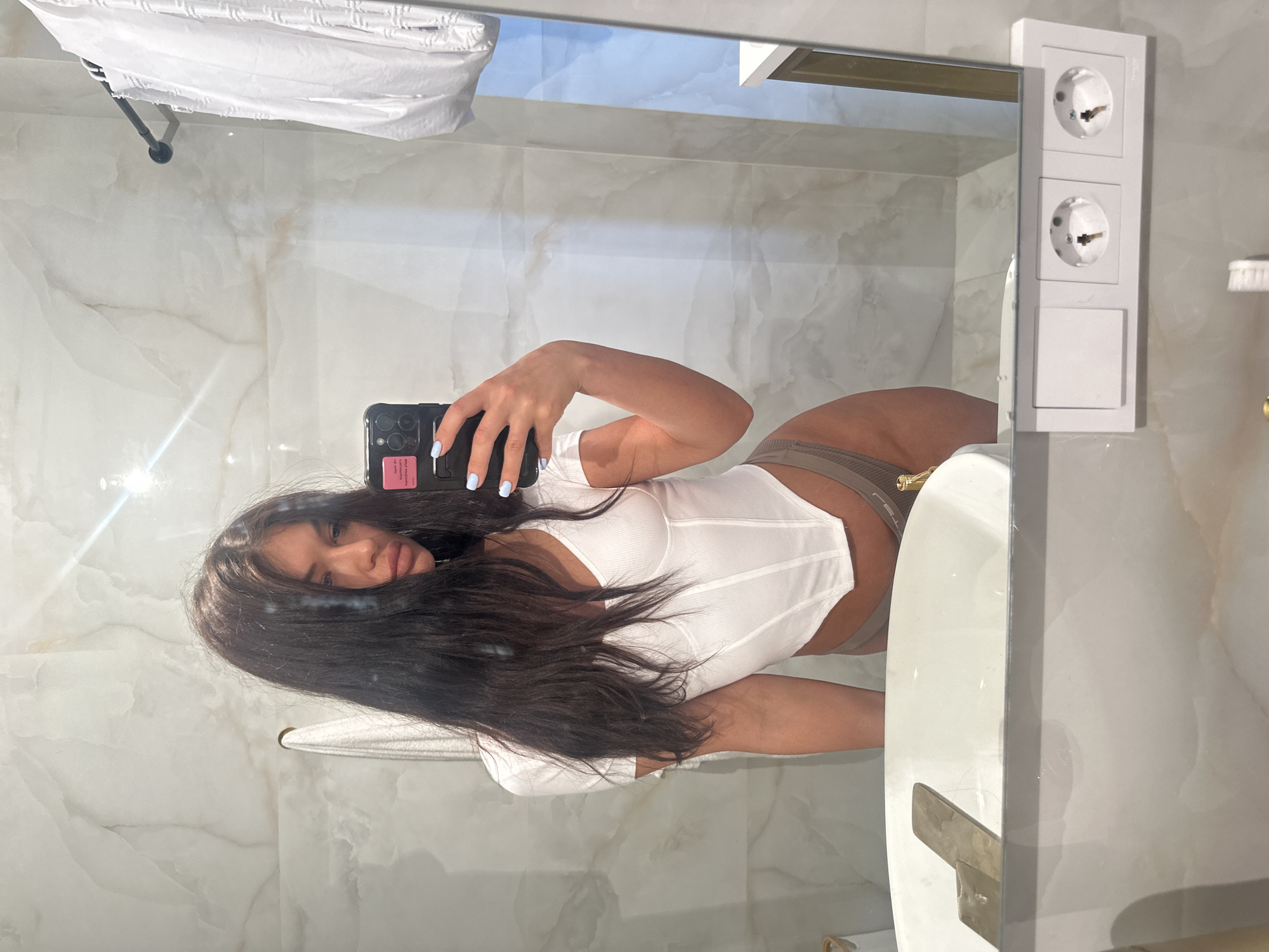 A young woman is captured in a mirror selfie taken in a modern bathroom, likely in Moscow, Russia, given the style of the electrical outlets. The image is oriented vertically, showing the woman's reflection in a large mirror. She has long, dark hair that frames her face and wears a white ribbed crop top with a corset-like design, paired with light grey or brown high-waisted underwear. Her left hand, adorned with light blue nail polish, holds a dark-colored smartphone that obscures her face partly, though her eyes are visible. A small pink sticker with unreadable text is visible on the back of the phone case.
The bathroom features light-colored, possibly marble-patterned, tiles on the walls, suggesting a clean and contemporary aesthetic. In the mirror's reflection, above the woman, a white towel hangs on a dark towel rail. To the right of the mirror, a white panel is mounted, containing two European-style electrical outlets stacked vertically, with a rectangular blank switch or cover plate underneath. A portion of a white sink or vanity is visible in the bottom right of the reflection. The scene is well-lit, indicating either bright natural light or effective artificial lighting.