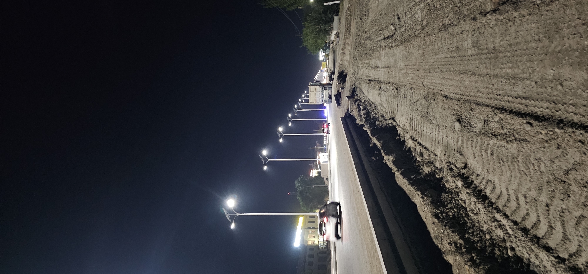 At nighttime, along a street undergoing construction in the city of Bukhara, Uzbekistan, a dual-lane road is illuminated by a series of streetlights. The road is bordered by a concrete barrier on the left and a construction area with tire tracks in the dirt on the right. Several vehicles are visible on the road: a light-colored sedan driving away from the camera on the left, another dark-colored vehicle in the distance on the right, and a large dump truck also on the right. Buildings with illuminated windows are present on the left side of the street, and trees are scattered throughout the scene. The sky is dark, indicating it is late at night.