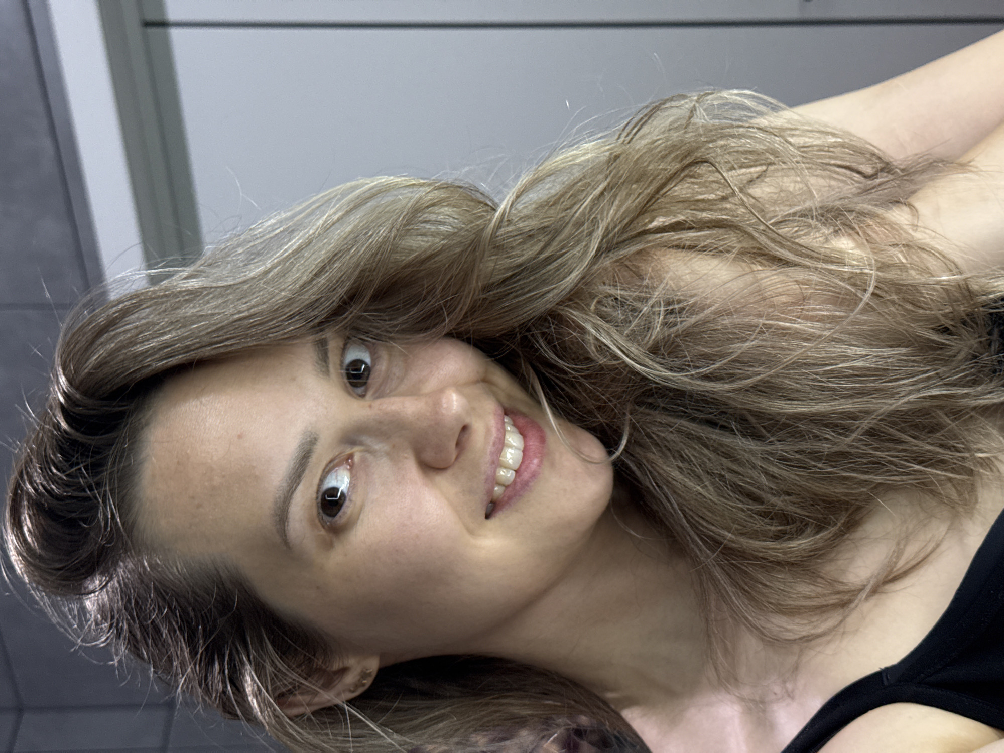 A portrait photograph captures a woman with long, wavy, light brown to blonde hair, looking directly at the camera with a broad, cheerful smile. Her eyes are wide open, conveying a playful or surprised expression. Her head is tilted slightly to her right, and her voluminous hair frames her face and flows around her shoulders. She has fair skin, and her white teeth are visible as she smiles. A black strap or top is visible on her right side. The background consists of a uniformly lit, neutral grey interior space, featuring subtle horizontal lines that suggest architectural elements like ceiling panels or wall divisions in a modern building or hallway. The lighting is even and artificial. No specific objects or text are visible in the image. The scene is set indoors, with no discernible weather or time of day cues. The provided context indicates the location is Chaikovsky, Russia, though no specific geographical markers are present in the visual.