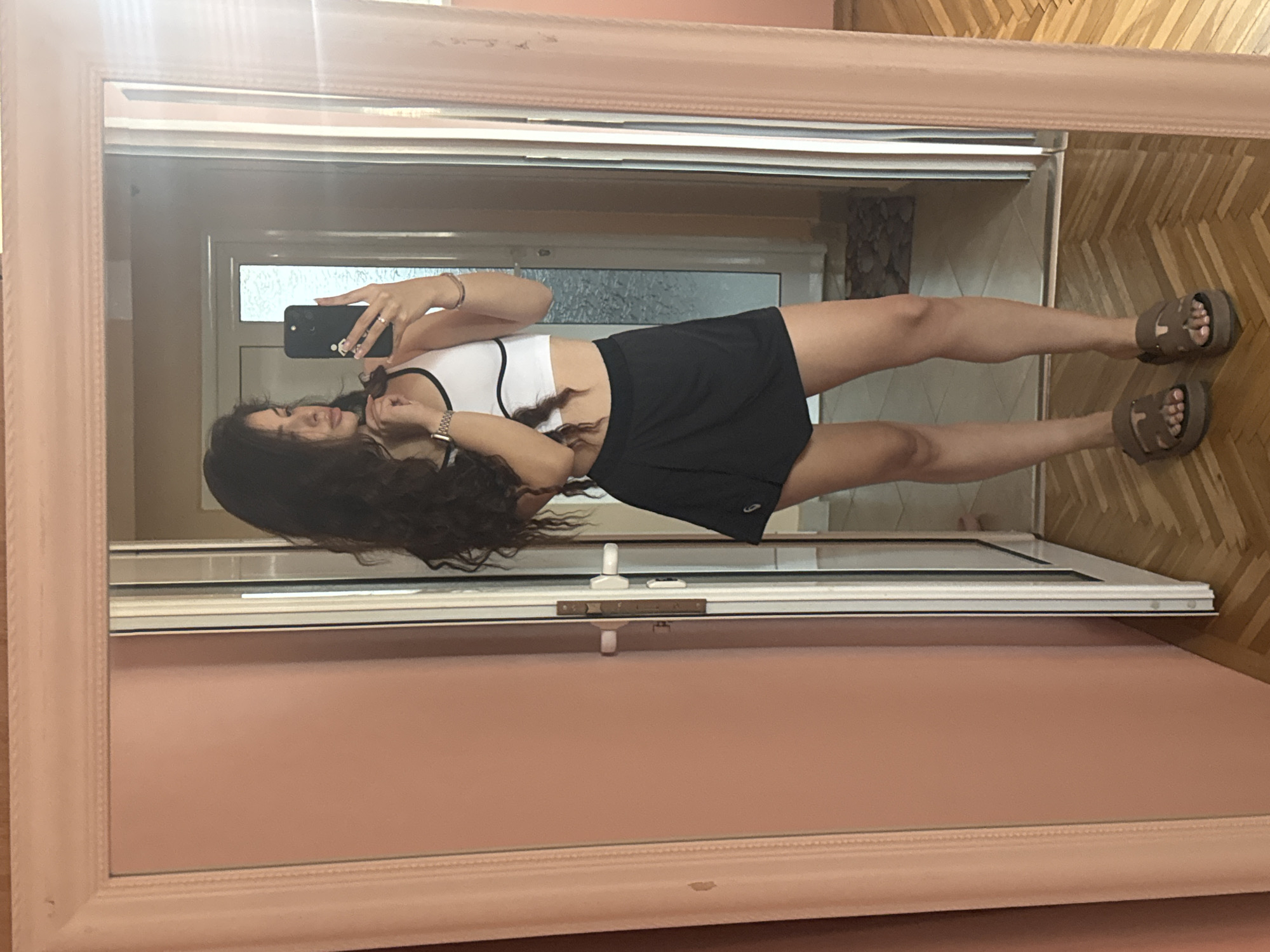A young woman is taking a full-length mirror selfie in a room. She is standing in front of a large mirror with a pink frame, which is leaning against a pale pink wall. She has long, dark, curly hair and is wearing a white sports bra with black trim, a black skort, and brown platform sandals. She is holding a black smartphone in her right hand, angled to capture her reflection. Her left hand is touching her neck. The background visible in the mirror shows a doorway, a glimpse of a hallway, and a wooden floor with a herringbone pattern. The scene appears to be an interior residential setting, potentially in Plovdiv, Bulgaria, given the style of the door and flooring, which are common in older Bulgarian apartments. There are no other people visible in the image.