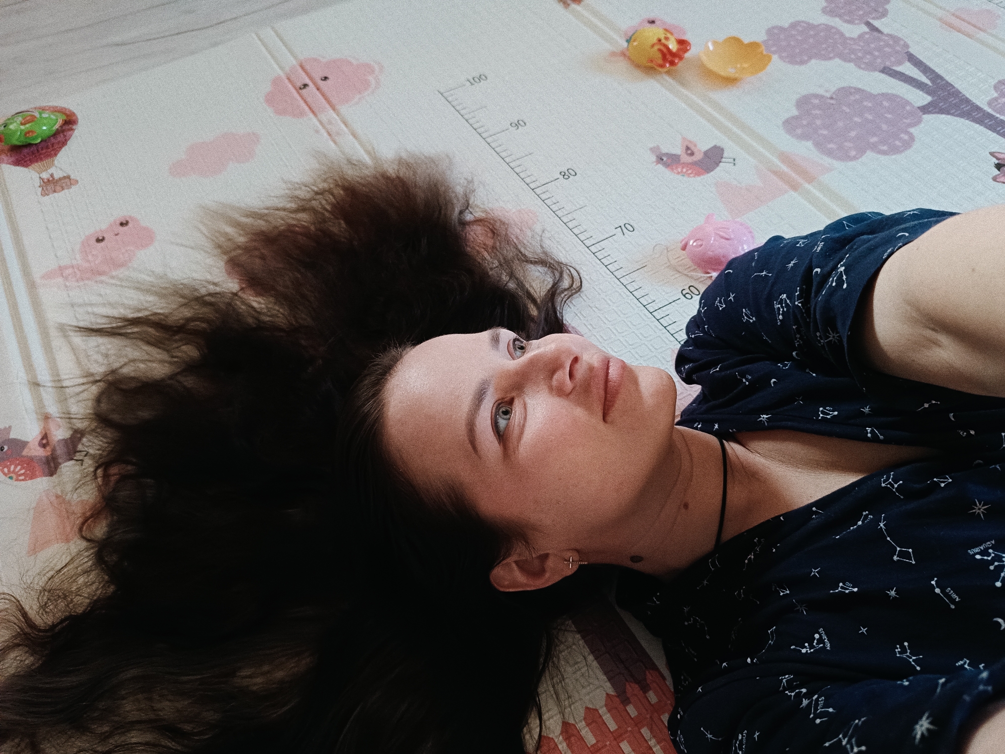A woman is lying on a play mat with a height chart printed on it. She has long, dark hair spread out around her head and is wearing a dark blue shirt with constellations printed on it. She is looking up and slightly to the left. There are several plastic toys scattered around her on the play mat. The location is Unknown, Unknown.