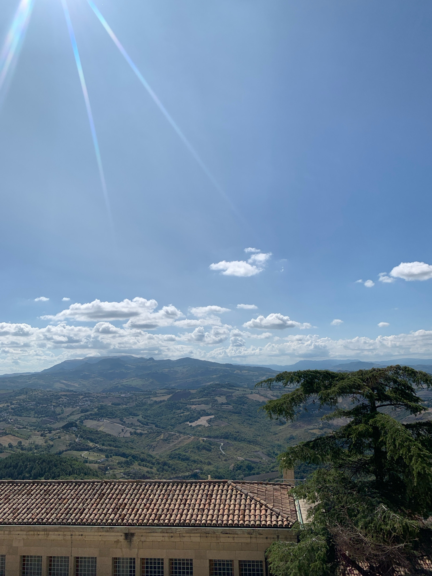 A telephoto shot captures a panoramic view of the City of San Marino, San Marino. The foreground is dominated by the terracotta tiled roof of a building, with a stone facade and multi-paned windows visible below. To the right, a large evergreen tree with dense foliage extends into the frame. The middle ground reveals rolling green hills dotted with smaller buildings and winding roads, leading to distant mountains. The sky above is a clear blue, interrupted by scattered white clouds. Sunlight streams from the upper left corner, creating lens flare and highlighting the cloud formations. The overall scene evokes a sense of tranquility and expansive natural beauty.