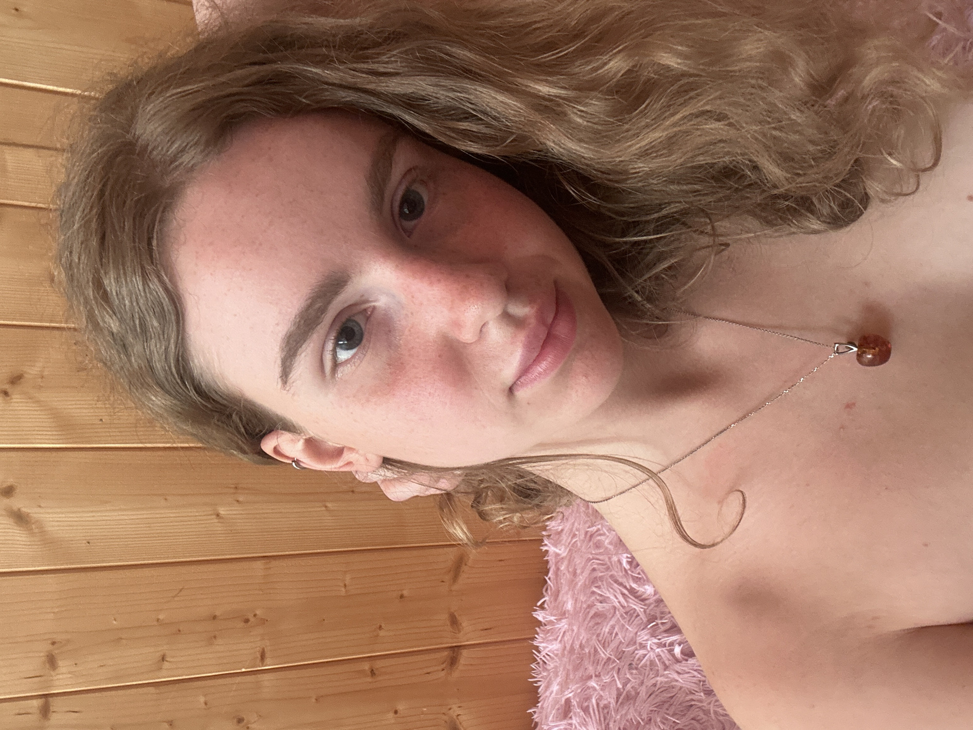 A close-up, slightly angled portrait captures a young woman with light brown, curly hair, likely in an indoor setting. Her head is tilted to the side, and she looks directly at the camera with a gentle, soft smile. Her fair skin shows a scattering of freckles across her nose and cheeks.
She is wearing a delicate silver-toned necklace with a small, amber-colored, tear-drop shaped pendant. A subtle hoop earring is visible in her left ear. Her shoulders are bare, suggesting she might be wearing a strapless garment or covered by a blanket. She appears to be lying on a fluffy pink blanket or fabric, parts of which are visible in the foreground.
The background consists of a wooden wall with horizontal planks, indicating a rustic or cozy interior. The lighting is bright and natural, suggesting it is daytime. The overall impression is one of peacefulness and comfort. No visible text is present in the image. The provided location context is Vasilyevo, Russia.