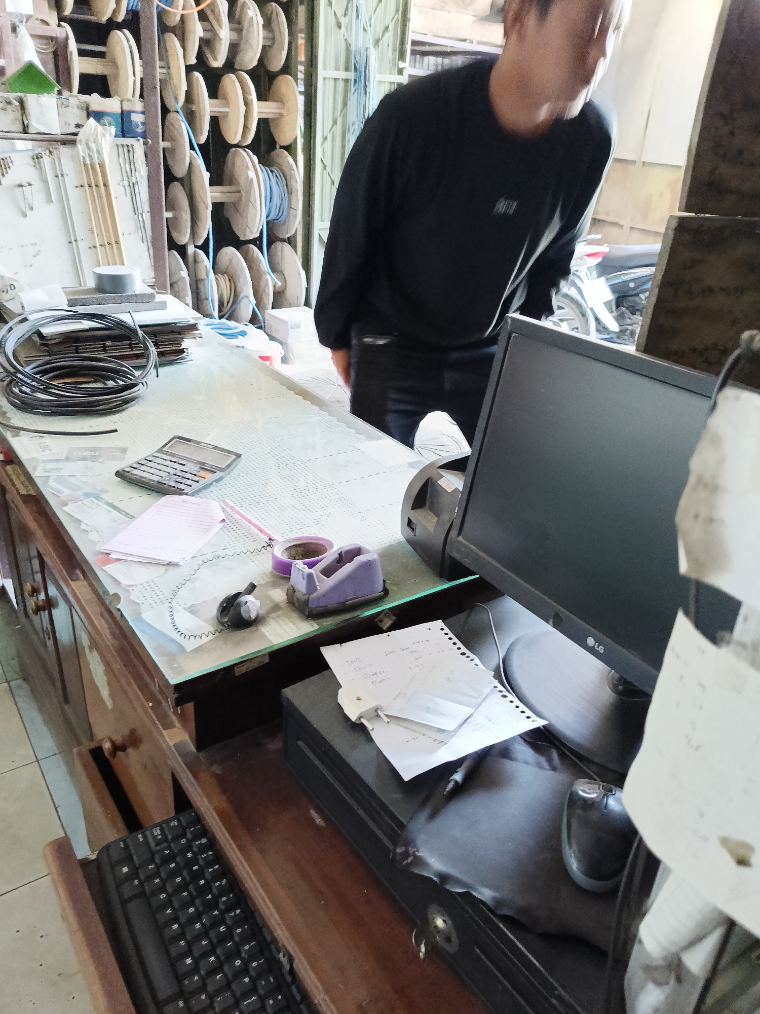The image depicts an indoor scene, likely a workshop or a store, located in Sukorini, Indonesia. A person, partially visible from the side and wearing a black long-sleeved shirt and dark pants, is bending over a wooden desk. The desk is cluttered with various items including a calculator, a notepad, a roll of purple tape with a dispenser, a small black handset, and a computer monitor. Behind the desk, an LG computer monitor is placed on a black computer tower. To the left of the desk, a keyboard is partially visible in an open drawer. The background features shelves stacked with spools of wire or cable, and some electrical equipment. A motorcycle is also partially visible in the background. The lighting suggests an indoor environment with overhead or natural light sources.