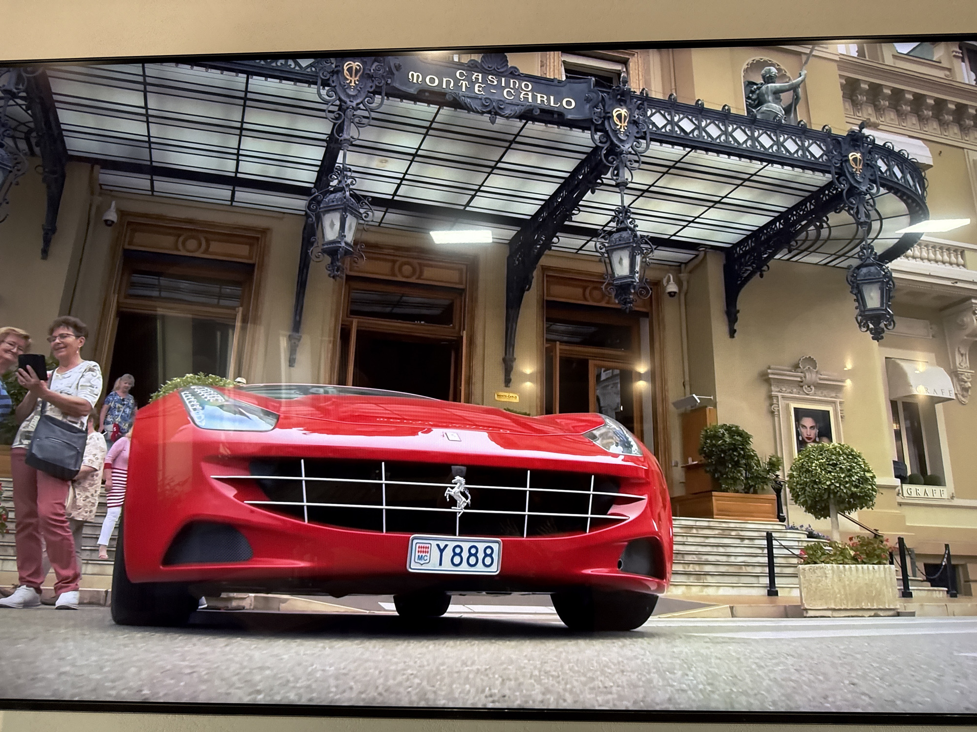 The image depicts a red Ferrari sports car parked in front of the entrance to the Casino de Monte-Carlo, a well-known landmark in Monaco. The casino features an ornate facade with a large canopy supported by decorative ironwork, hanging lanterns, and a sign that reads "CASINO MONTE-CARLO". Above the canopy, a statue of a figure holding a spear is visible.
To the left of the car, two women are standing on the steps leading into the casino. One woman, wearing pink trousers and a white patterned top, is holding a tablet. The other woman, in a blue patterned top, is standing behind her. Another person in a pink and white striped shirt is visible further up the steps.
The car's license plate is visible, displaying "Y888" with what appears to be the Monaco insignia. The scene suggests a luxurious setting, possibly a film scene or a high-profile event, given the presence of the sports car and the iconic location. There is no indication that this scene is located in Dnipro, Ukraine.