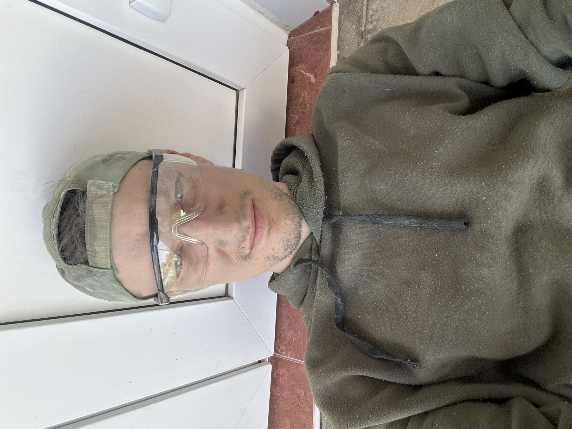 In Makiv, Ukraine, a selfie captures a man from the chest up, wearing a camouflage baseball cap and clear safety glasses. He is dressed in a dark olive green hoodie with a drawstring. The background shows white vertical window frames and a reddish-brown tiled floor. The man's face is partially obscured by the glasses, and his expression is neutral. The hoodie appears worn and has small white specks scattered across its surface, suggesting dust or lint. The lighting suggests an outdoor or well-lit indoor environment.