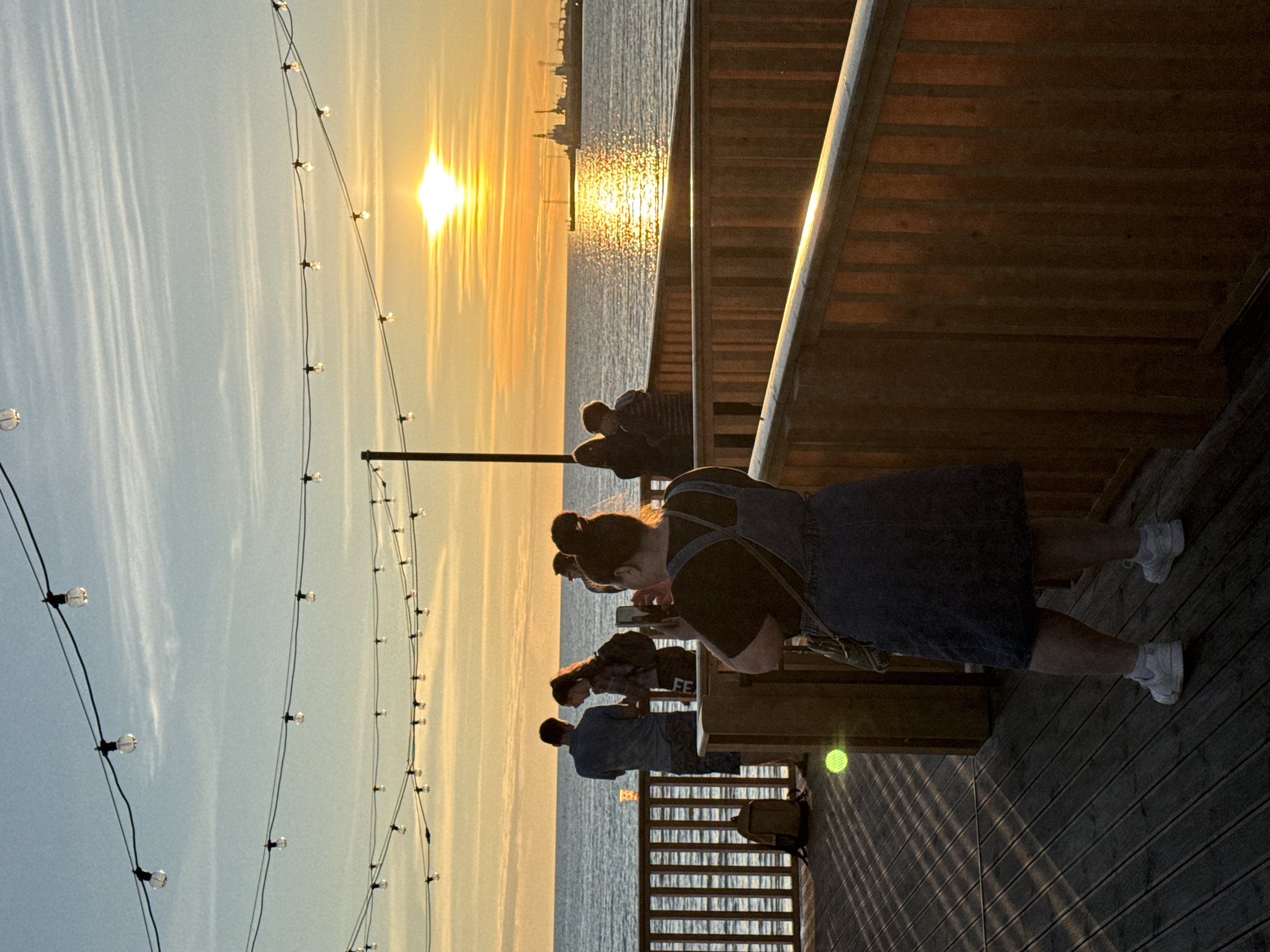 An outdoor shot captures a sunset over a body of water, possibly a harbor, with a low wooden railing in the foreground and mid-ground. The sky is a blend of blue and orange, with wispy clouds. String lights with exposed bulbs are strung across the top of the frame.
In the foreground, a woman with her hair in a bun is standing, facing away from the camera and towards the water, holding a phone to take a picture. She is wearing a denim jumpsuit over a dark shirt and white sneakers. To her left, on the wooden deck, a brown backpack is leaning against the railing.
Further back, near the railing, a group of people are gathered. Two figures, a man in a blue shirt and shorts and a woman with a backpack, are standing closest to the water, looking out. Behind them, other figures are partially visible.
On the water, a large industrial structure or ship is visible in the distance, silhouetted against the bright setting sun. The sun's reflection creates a shimmering path across the water.
The wooden deck has a slatted pattern, and shadows from the railing and overhead lights create a striped effect. The overall atmosphere is that of a relaxed evening gathering by the water. The specific location within Tallinn, Estonia, cannot be definitively determined from the visual information provided.