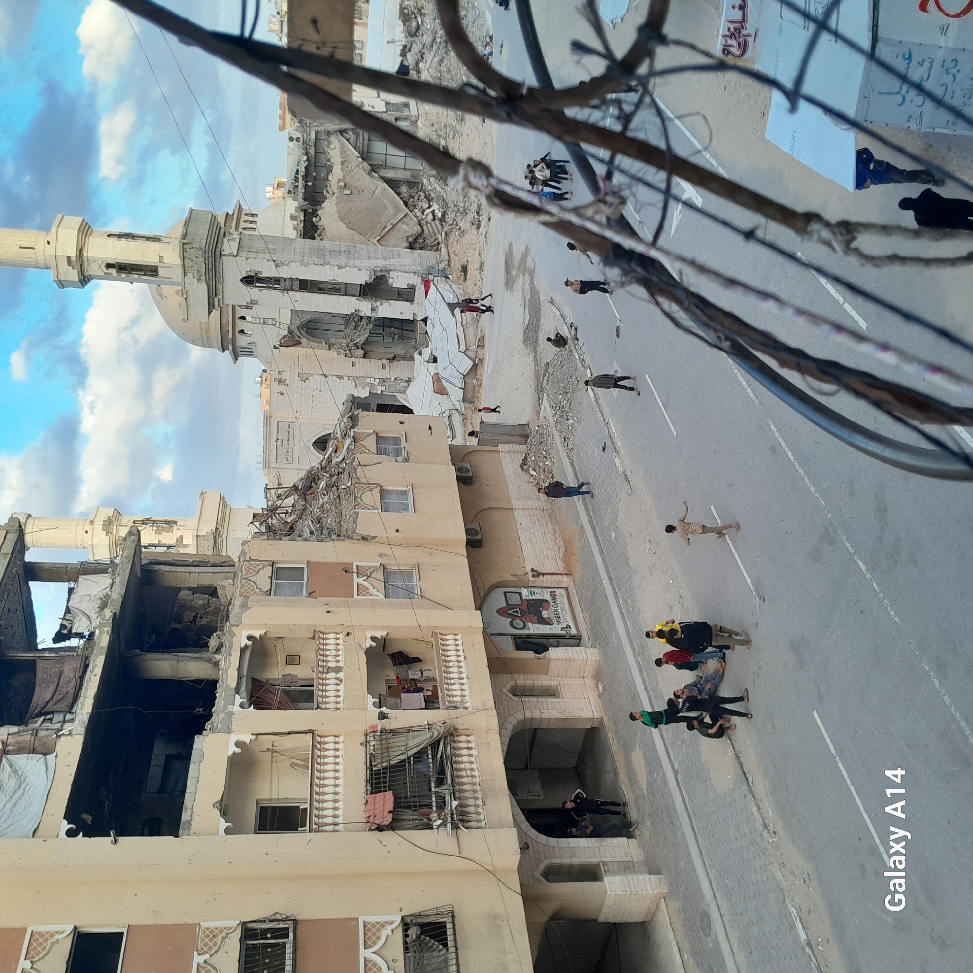 The image shows a street scene in a city, with damaged buildings including a mosque, indicating potential conflict or disaster. Several people are visible on the street. A banner with "YASEEN GAMES" can be spotted on one of the buildings. Also, "Galaxy A14" is written on the bottom right.