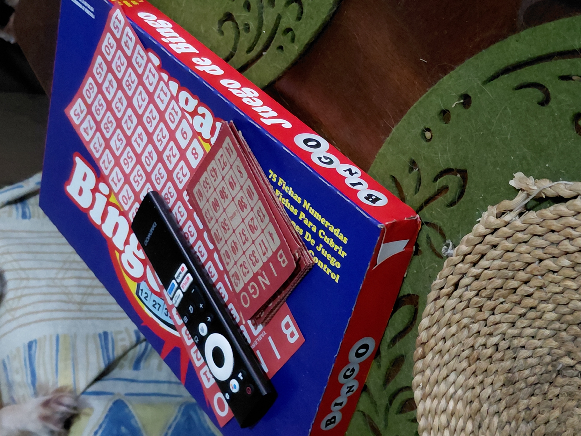 The image shows a Bingo game box and its components. The box is blue, red, and white, with "Bingo Juego de Bingo" printed on it. Stacks of Bingo cards and 75 chips are visible. A black remote control is placed on top of the Bingo box. The setting is indoors, with a patterned cushion or fabric to the left and a woven placemat or basket to the right. Part of an animal, possibly a pet, is seen on the left. The location, Santa Ana, Venezuela, is not discernible from the visual data.