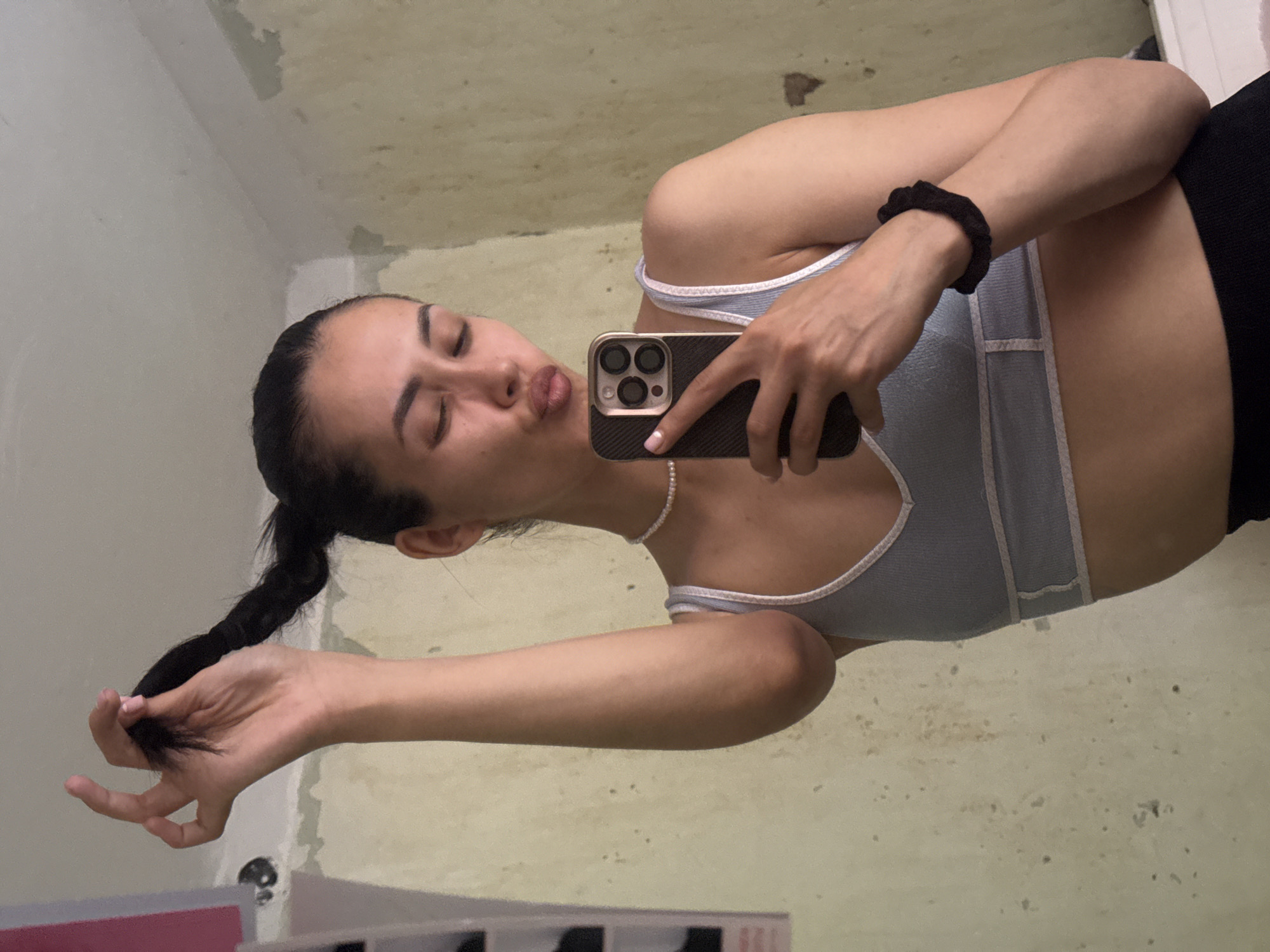 A young woman, possibly in her late teens or early twenties, is taking a selfie in what appears to be a dimly lit room. The room's walls are a light, uneven color, suggesting unfinished or weathered plaster, with some darker splotches scattered across them. The ceiling also shows similar textures. The lighting is dim, casting a yellowish hue over the scene, hinting at an indoor setting, possibly a bathroom or a poorly lit bedroom, at night or in a windowless space.
The woman has dark hair pulled back into a low ponytail and is wearing a light blue tank top with white trim. She is holding a smartphone in her right hand, angled to capture her reflection. Her eyes are closed, and her lips are pursed in a kissy-face gesture, suggesting a playful or flirtatious mood. On her left wrist, she wears a black scrunchie. Her left hand is holding a few strands of her braided hair. A delicate pearl necklace is visible around her neck.
The camera is angled such that the woman's reflection is prominent, with a portion of her torso and arm visible. The background, seen in the reflection, is the same wall and ceiling material. The overall impression is a casual, informal moment captured in a somewhat austere environment. There is no visible text in the image.