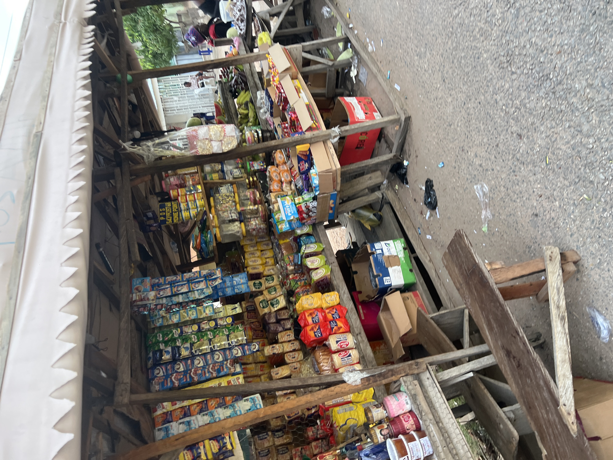 This image depicts an informal outdoor market stall operating on a paved surface in Jalingo, Nigeria. The stall, constructed from unpainted wooden planks and poles, features a white fabric awning with a serrated lower edge. Various packaged goods are displayed on multiple wooden shelves and hanging from strings within the stall.
On the left, shelves hold tubs of what appear to be cosmetics or food items, alongside various small packaged snacks. A yellow sign displaying "POS 6439335768 MONEY POINT" is affixed to a wooden support. The central part of the stall showcases numerous sachets of products hanging from the structure, alongside stacks of packaged biscuits and other dry goods in various colorful wrappers. Towards the right, additional shelves contain more packaged items, including boxes of sweets and biscuits. Below the shelves, cardboard boxes are utilized for storage.
A section to the right of the main stall area displays fresh produce, including bananas and watermelons, on a wooden table. Partially obscured individuals are visible behind this section, with one person wearing a purple headwrap and red attire attending to the produce. In the background, several people are present; one individual is seated on a white chair near a building with a white gate.
The ground surrounding the stall is an asphalt or concrete surface, visibly scattered with plastic bags, wrappers, and other debris, indicative of an active market environment. A simple wooden bench made of rough planks rests on the left side in the foreground.