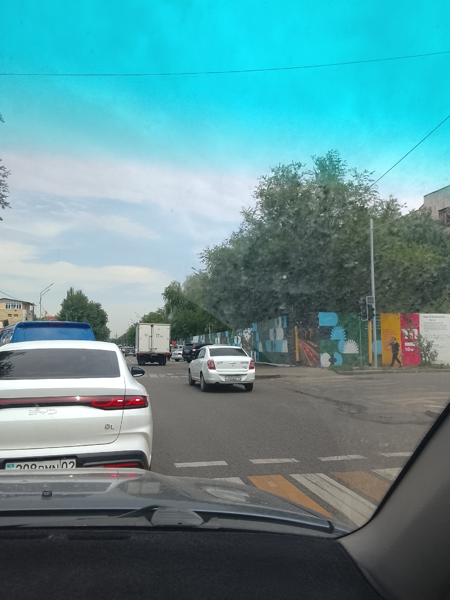 The image captures a street scene in Almaty, Kazakhstan, viewed from inside a vehicle, likely at a traffic intersection. The sky is a vivid turquoise, possibly due to tinted glass or atmospheric conditions. The road is moderately busy with vehicles, including a white BYD sedan in the foreground, a white Chevrolet sedan, a white truck, and a blue van. A pedestrian is visible walking on the sidewalk near a construction site adorned with colorful murals. The environment includes mature trees lining the street and buildings in the background. The presence of a crosswalk and traffic signals indicates this is a public thoroughfare.
