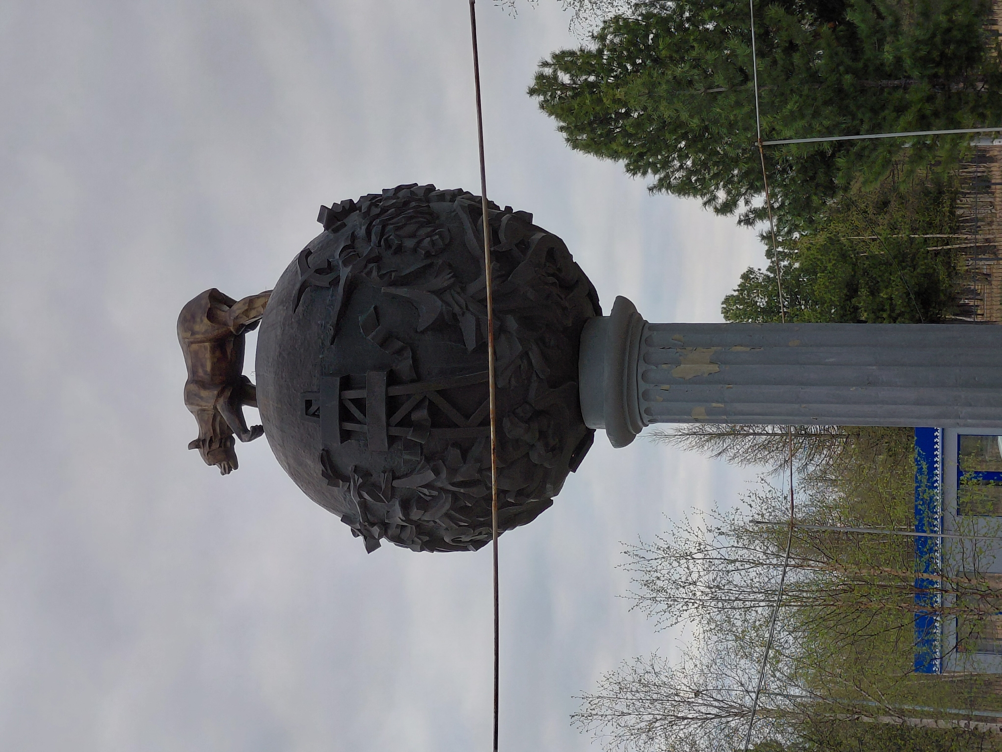 Here's a description of the image:
The image shows a statue in the city of Nadym, Russia. The statue consists of a bronze figure of a wolverine on top of a dark-colored sphere, which is decorated with carvings. The sphere is supported by a grey, fluted column. The statue is set against a backdrop of trees and an overcast sky. A wire or cable crosses the frame. A partial view of a blue and white building can be seen in the background.