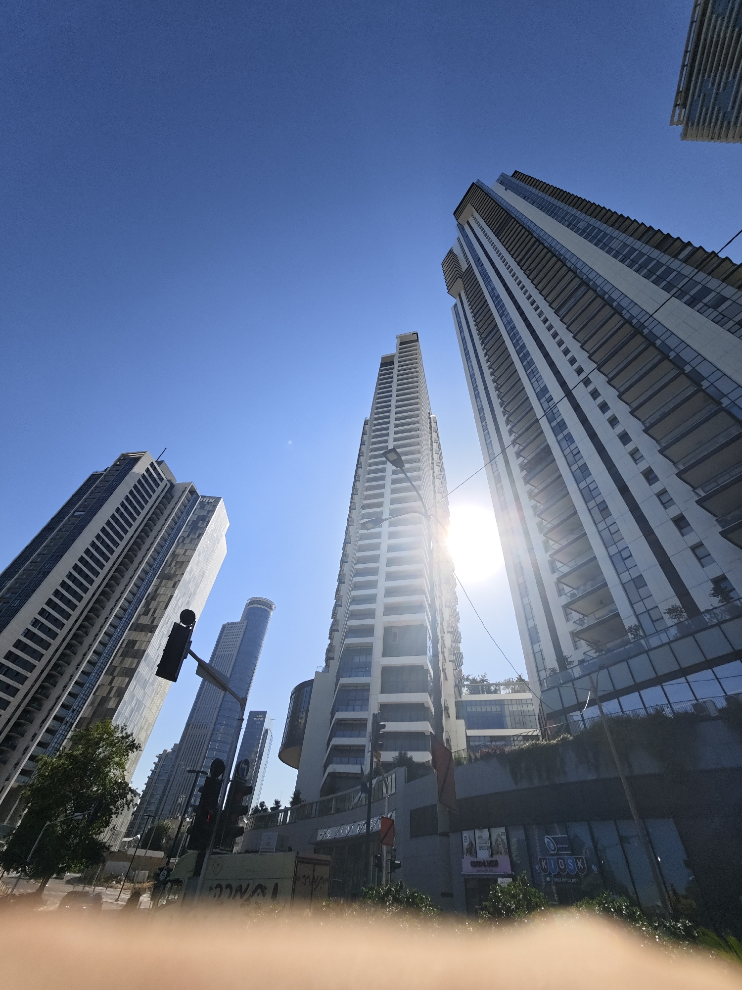 A low-angle, wide-shot captures a cluster of modern high-rise buildings under a clear blue sky, likely in the city of Givatayim, Israel. The sun shines brightly, creating lens flares and casting glares on the buildings' glass facades. Multiple skyscrapers, characterized by their sleek designs and balconies, dominate the frame, angled towards the camera. Among them, a traffic light is visible, indicating an urban street scene. Some lower structures and trees are present at the base of the buildings, suggesting a commercial or residential area within the city. The foreground is blurred with what appears to be a person's fingers, obscuring the very bottom of the image.
