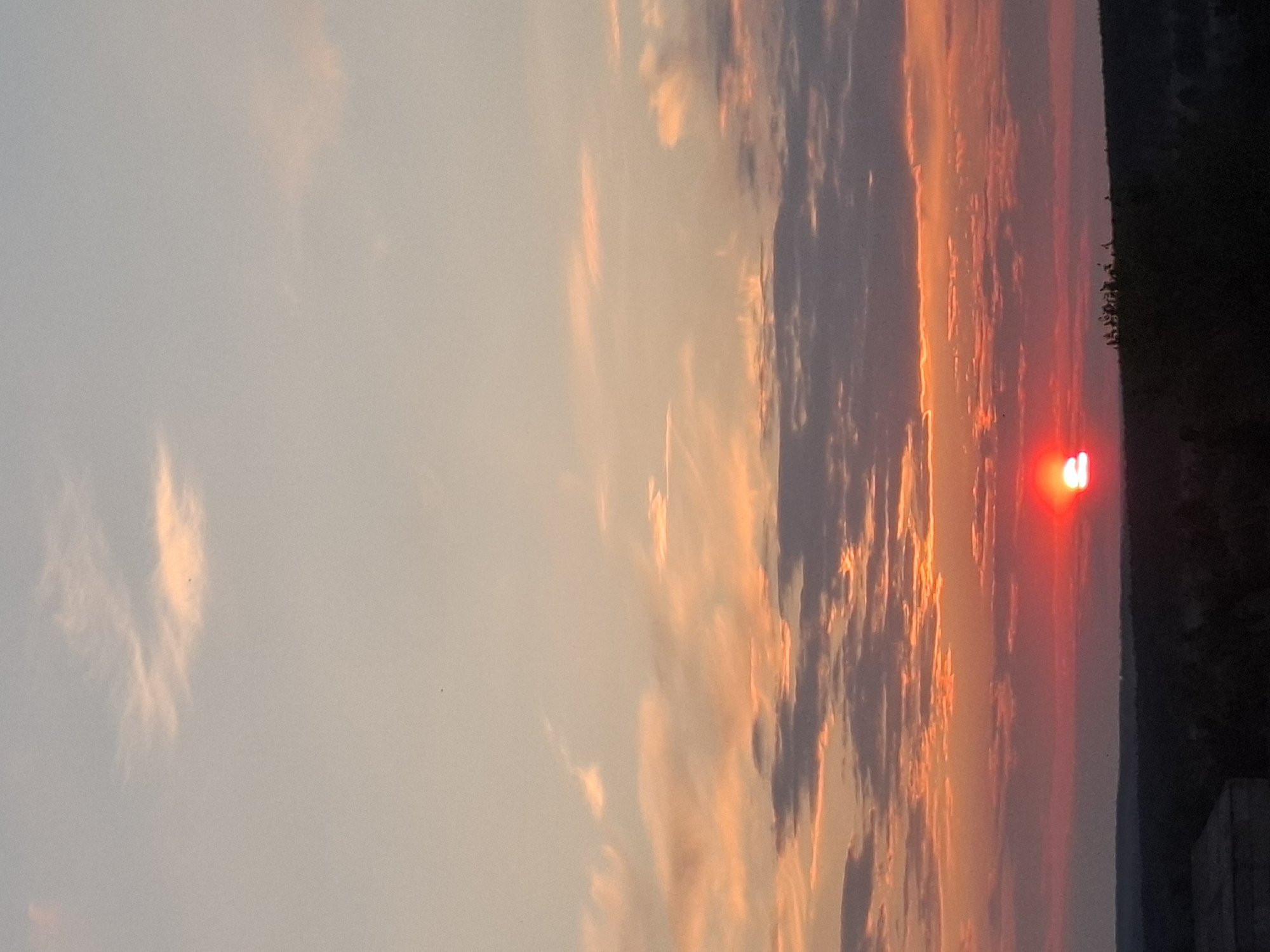 The image shows a sunset in what is possibly Khlevnoye, Russia. The sky is filled with clouds tinted orange and pink by the setting sun. In the lower right corner, there is a dark area with some trees, potentially indicating a wooded area or the edge of a settlement. The sun itself is visible as a bright red object near the horizon. The orientation of the image is sideways.