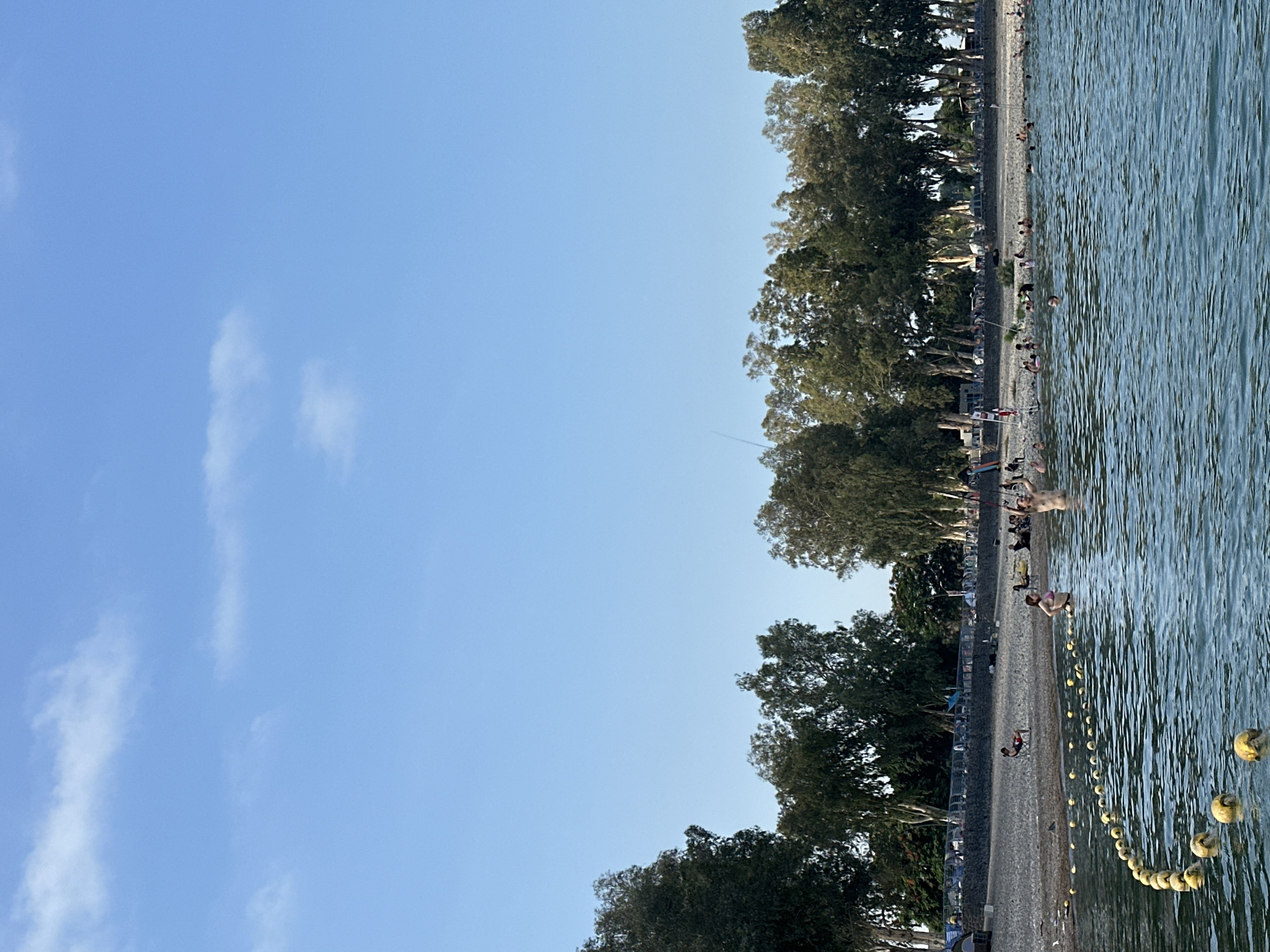 The image depicts a busy beach scene at the Sea of Galilee in Tiberias, Israel. The foreground shows the clear blue water with a yellow buoy line delineating a swimming area. Several people are visible in the water, some swimming, others standing or relaxing. The beach behind them is a mixture of pebbles and sand, with more people seated and lying down, enjoying the sun. A line of trees borders the beach, providing a backdrop of green foliage. The sky is a bright, clear blue with a few wispy clouds, suggesting a pleasant day. The overall impression is one of a popular recreational spot, with many individuals engaging in various beach activities.