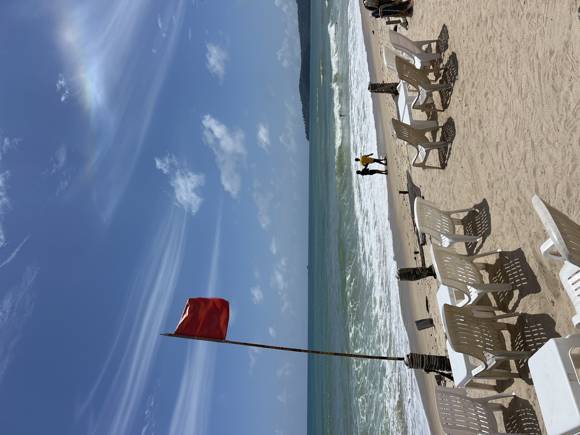 A red flag flies on a pole on a sandy beach in Choeng Thale, Thailand, near the ocean. The flag is blowing in the wind. The ocean has moderate waves and is a turquoise color. There are several white plastic chairs and tables set up on the beach, facing the water. Two people, one wearing a black shirt and the other a yellow shirt, are walking along the shoreline. The sky is blue with some scattered white clouds. There is a faint rainbow visible in the sky. The scene is bright and sunny.