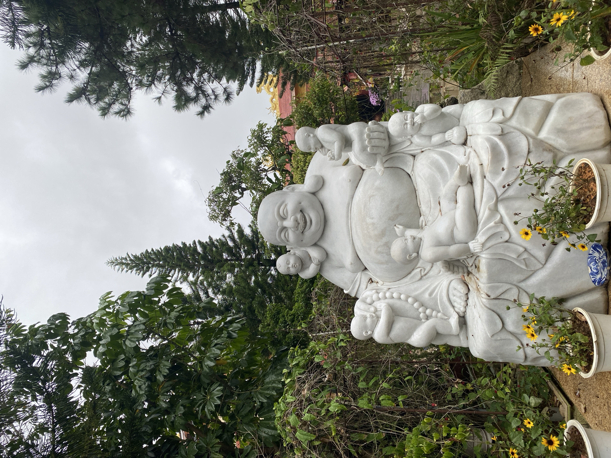 A large, white marble statue of a laughing Buddha is prominently displayed outdoors, surrounded by lush greenery and trees. The Buddha is seated on a sculpted base, adorned with several smaller figures of children, some of whom are perched on the Buddha's lap and shoulders, and others are interacting with him. The setting appears to be a garden or temple courtyard, with a hint of traditional Vietnamese architecture visible in the background, including a gilded roof with dragon motifs. The sky is overcast, suggesting a cloudy day. The statue is positioned in what is identified as the Lam Vien Square area of Dalat, Vietnam, a popular tourist destination known for its serene landscapes and cultural attractions. The foreground features potted plants with yellow flowers, adding a touch of color to the scene.