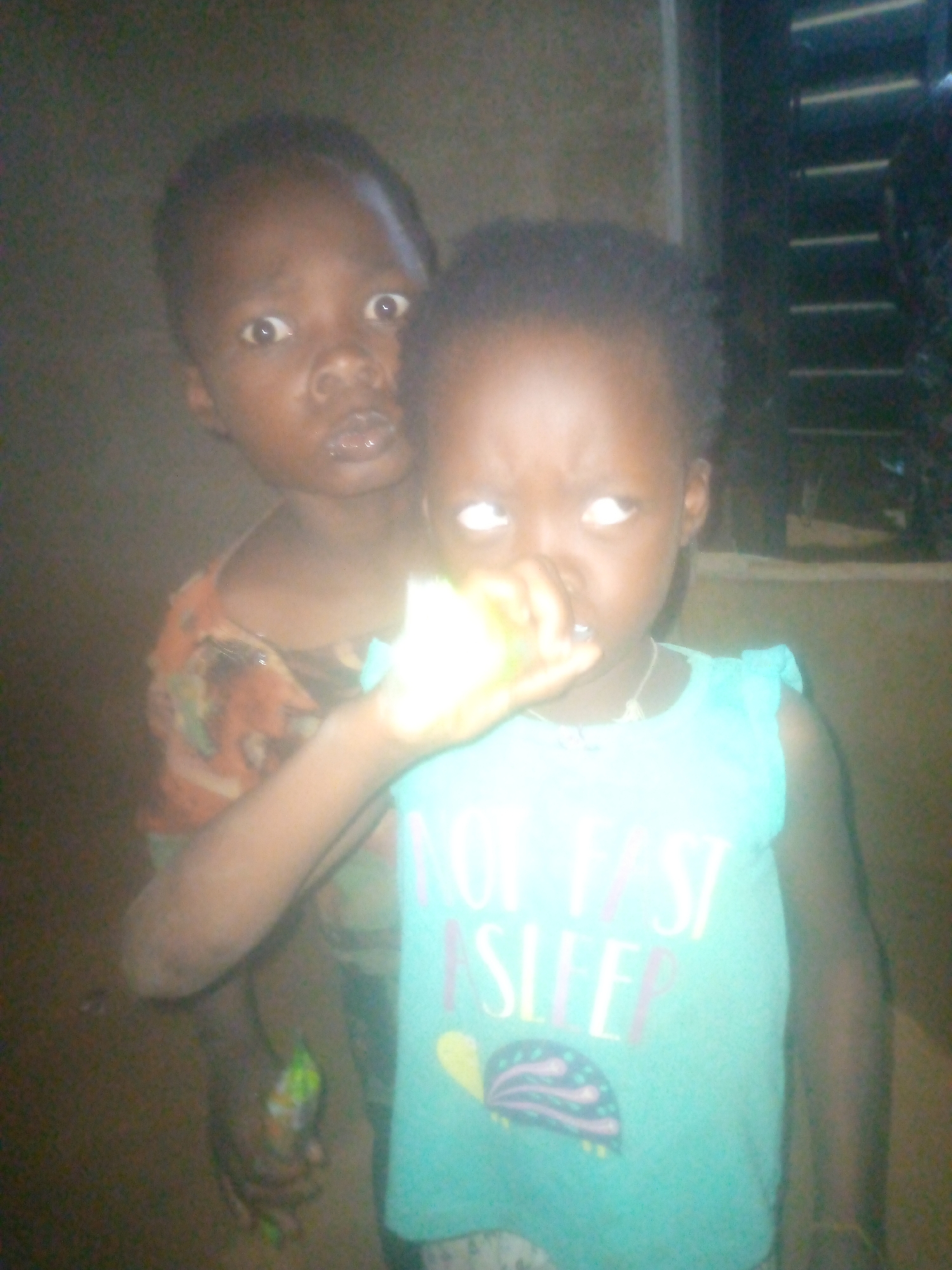 Two young children stand close together indoors. The child in front wears a light blue sleeveless shirt with the words "NOT FAST SLEEP" printed on it and a colorful turtle graphic. They are holding something small and indistinct in their hand near their face. The child behind them wears a patterned shirt in shades of orange and green. Their facial expressions are distinct, with the child behind appearing surprised or concerned. The background includes a window with horizontal slats. The location is Unknown, Unknown.