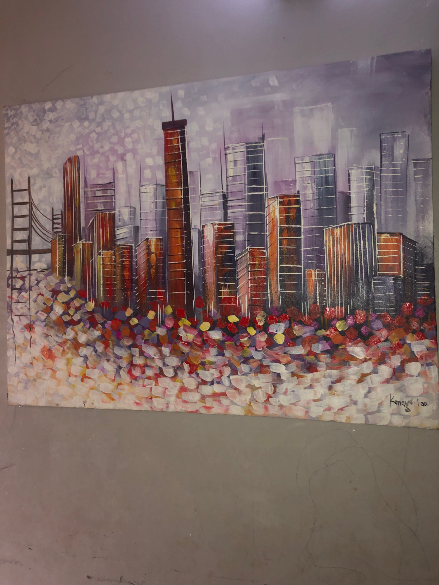 The image displays an abstract painting of a cityscape, likely depicting skyscrapers and a bridge. The foreground is filled with a vibrant, textured mosaic of colorful brushstrokes in shades of white, pink, red, orange, yellow, and purple, possibly representing flowers or reflective light. Behind this, a cluster of stylized buildings rises, with vertical lines suggesting windows and structural details. Some buildings are rendered in warm tones of orange and brown, while others are in cooler shades of gray and purple. A bridge structure with ladder-like supports is visible on the left side of the painting. The sky above is a hazy blend of purple and white, with textured brushstrokes creating a cloudy effect. The signature "Kangyo Lau 2012" is present in the lower right corner of the painting. The artwork is hung on a plain gray wall, with some faint pencil markings visible on the wall below the painting. There are no people visible in the image. The painting does not depict any specific landmarks or characteristics that would allow for identification of the city of Minna, Nigeria.