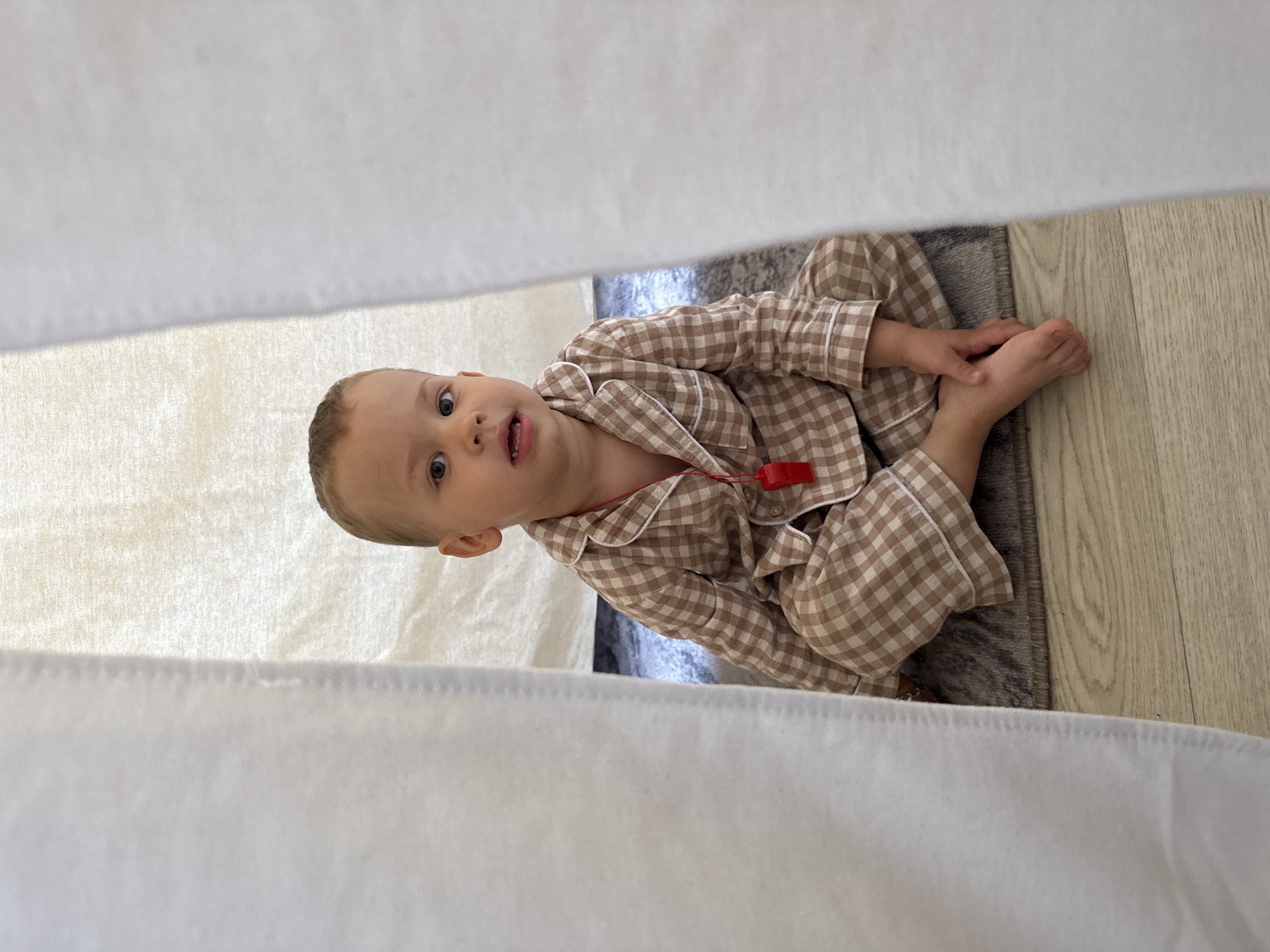 A young child wearing brown and white checkered pajamas is lying on the floor, partly on a gray rug, with bare feet visible. The child is positioned between two white fabric surfaces, suggesting a confined space like a play tent. A red whistle is visible around the child's neck. The floor is light wood-toned. The location appears to be indoors. Without additional information, determining the precise location in Rusanovo, Russia is impossible.