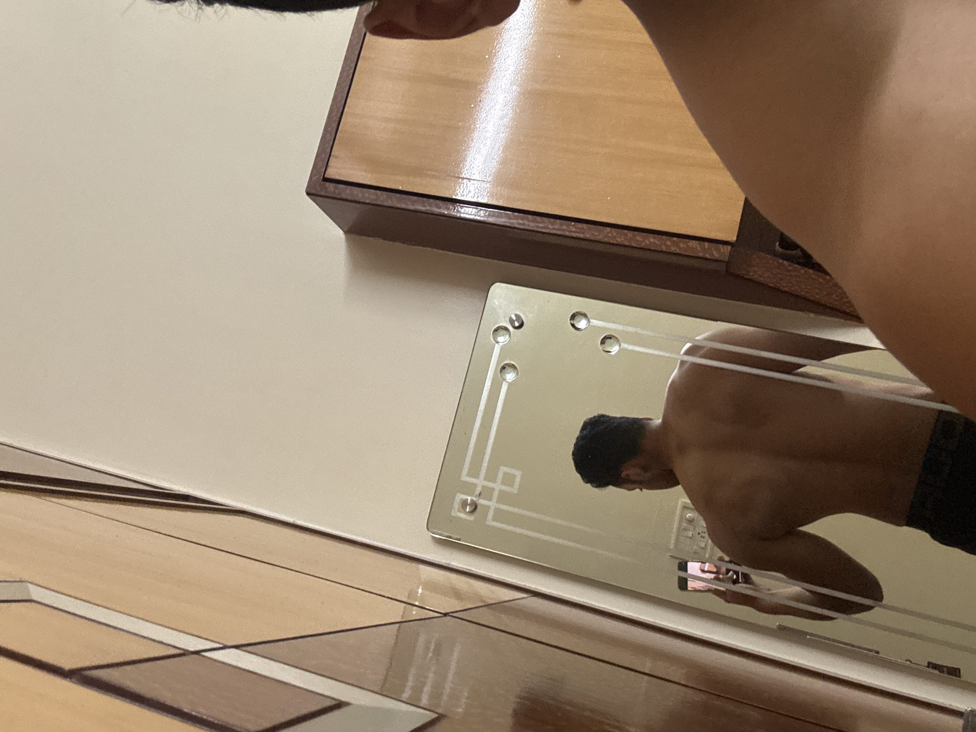 The image shows a partial view of a room's corner. A person is reflected in a small, decorated mirror mounted on the wall. The reflection shows a torso, with black hair and dark shorts. The person is holding a mobile phone, presumably taking the photograph. The wall is painted in a neutral color and has a wooden cabinet fixed to it. The floor is tiled with a geometric pattern of light and dark brown shades. There is no definitive indication of the location being Kolkata, India in the image.