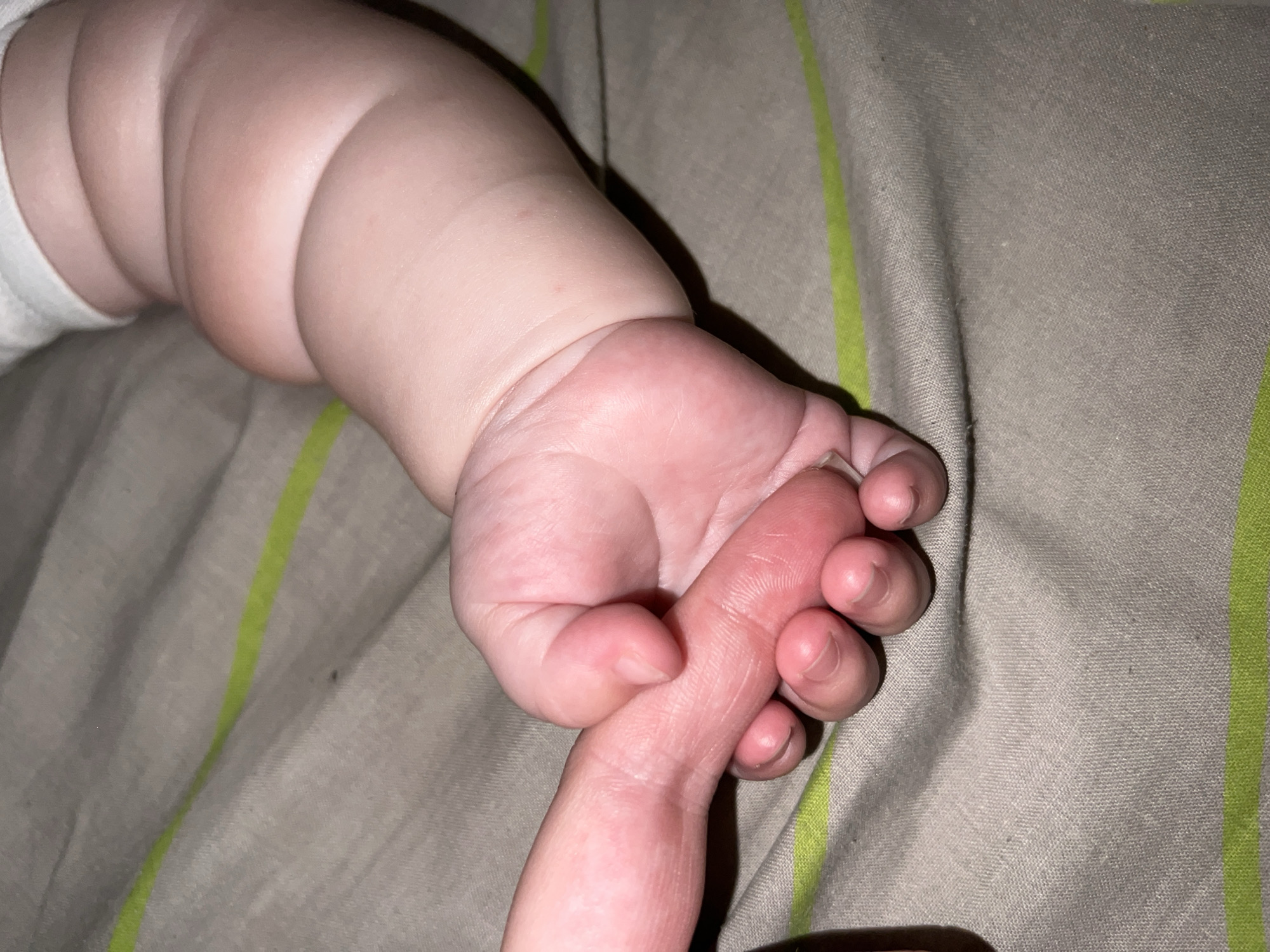 Two hands, one adult and one infant, are clasped together. The infant's hand is chubby and pink, with visible creases on the arm. The adult's hand is also visible, its fingers interlaced with the infant's. Both hands are resting on a beige fabric surface with thin green stripes. The lighting is dim, and the background is out of focus. The scene appears to be indoors, possibly in a home environment. The location is Unknown, Unknown.