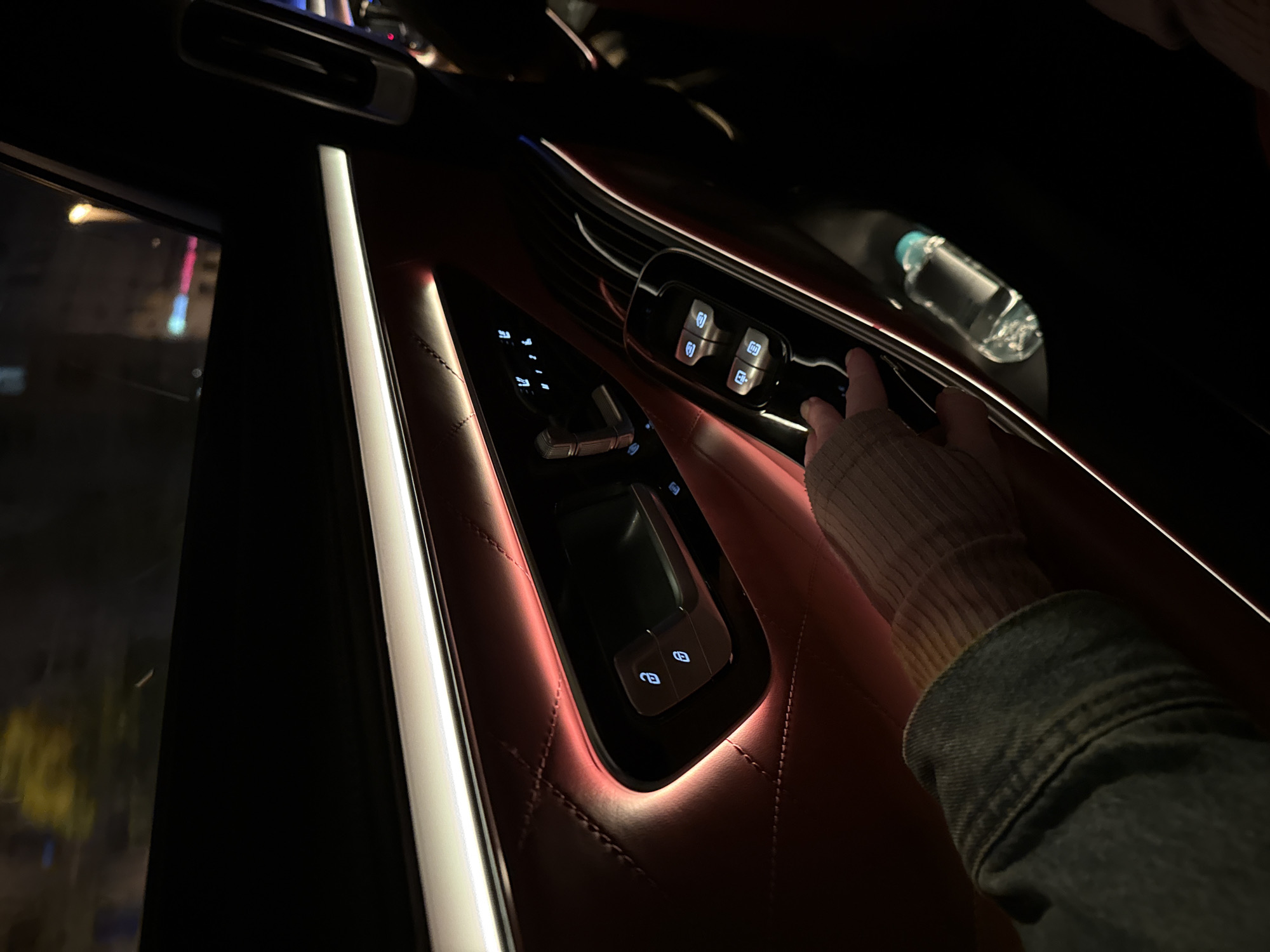 The image shows the interior of a car at night. The focus is on the driver's side door panel, which is upholstered in maroon quilted leather with white trim. A hand, wearing a ribbed sweater and a denim jacket, is operating the window controls. The buttons are illuminated in blue light. A water bottle is visible on the upper part of the door. Through the window, blurred lights suggest an urban environment. The city location is not discernible in this image.