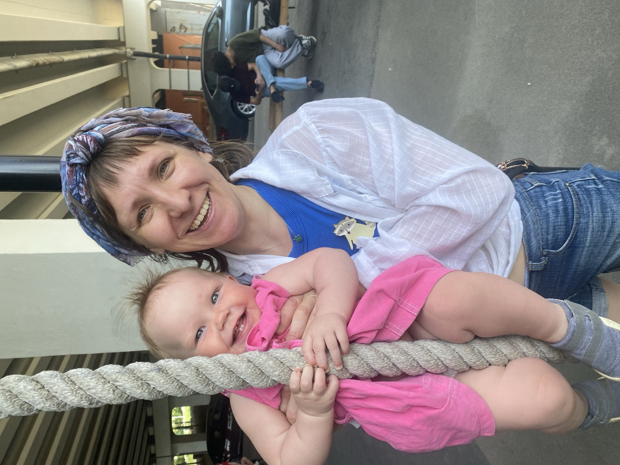 The image shows a woman and a baby posing with a rope structure, likely on a playground. The woman is smiling and wearing a patterned headscarf, a white shirt over a blue top, and denim shorts. The baby, dressed in pink, is also smiling and holding onto the rope.
In the background, blurred, there is a vehicle and two people interacting near it. The environment appears to be an urban setting, possibly a park area, under a structure that may be a bridge or an elevated roadway.
There is no direct evidence in the image to confirm its location in Yekaterinburg, Russia. The scene is an outdoor setting, where a woman holds a baby in front of a thick rope.