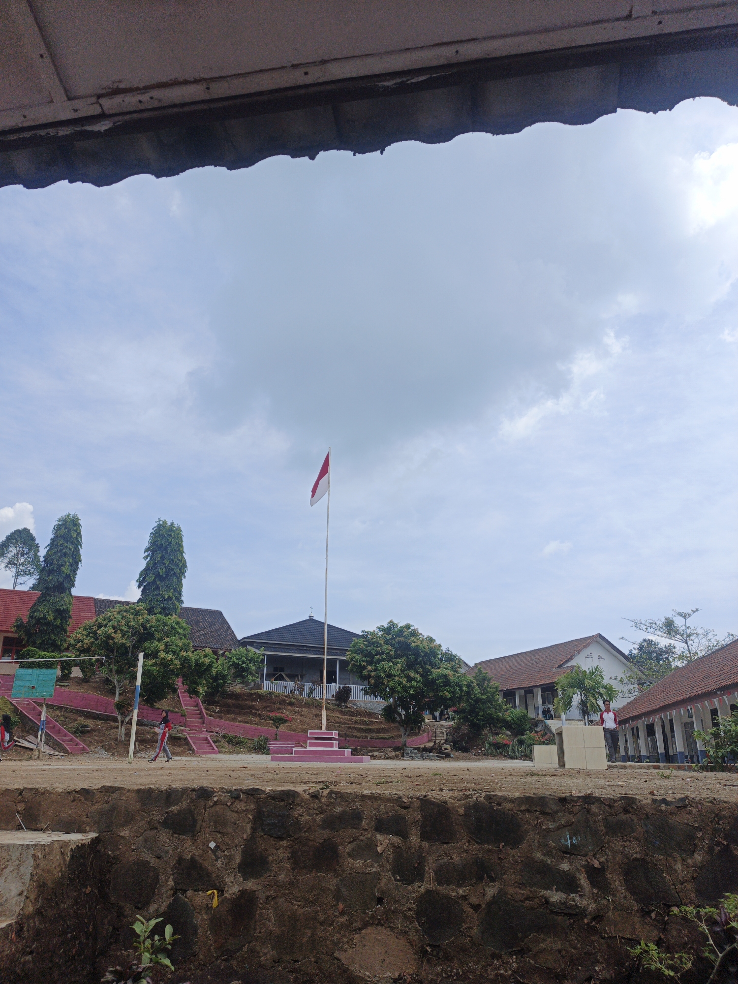 The image depicts an outdoor scene at what appears to be a school in Tanjung Setia, Indonesia, under a cloudy sky. In the foreground, a low wall constructed of dark, rough-hewn stones forms a barrier. Beyond the wall, a dirt path or open area is visible, with a few scattered green plants.
Dominating the mid-ground is a red and white Indonesian flag flying from a tall flagpole on the right side of a raised platform with tiered red steps. Behind the flag and platform, several buildings with terracotta tile roofs are visible. Some of these buildings have open-air verandas with white pillars. Trees, including tall, slender evergreen trees and smaller leafy trees, are interspersed among the buildings. A set of pink stairs with dark steps leads up a grassy embankment on the left side of the image.
Two individuals are present in the scene. On the left, a person wearing a dark headscarf and a red and white athletic outfit is walking across the open area, facing towards the right. On the far right of the mid-ground, a person wearing a white shirt and red vest is standing near a building. The overall impression is one of a school campus, possibly during a break or the school day. The sky is overcast with diffused light, suggesting it is daytime.