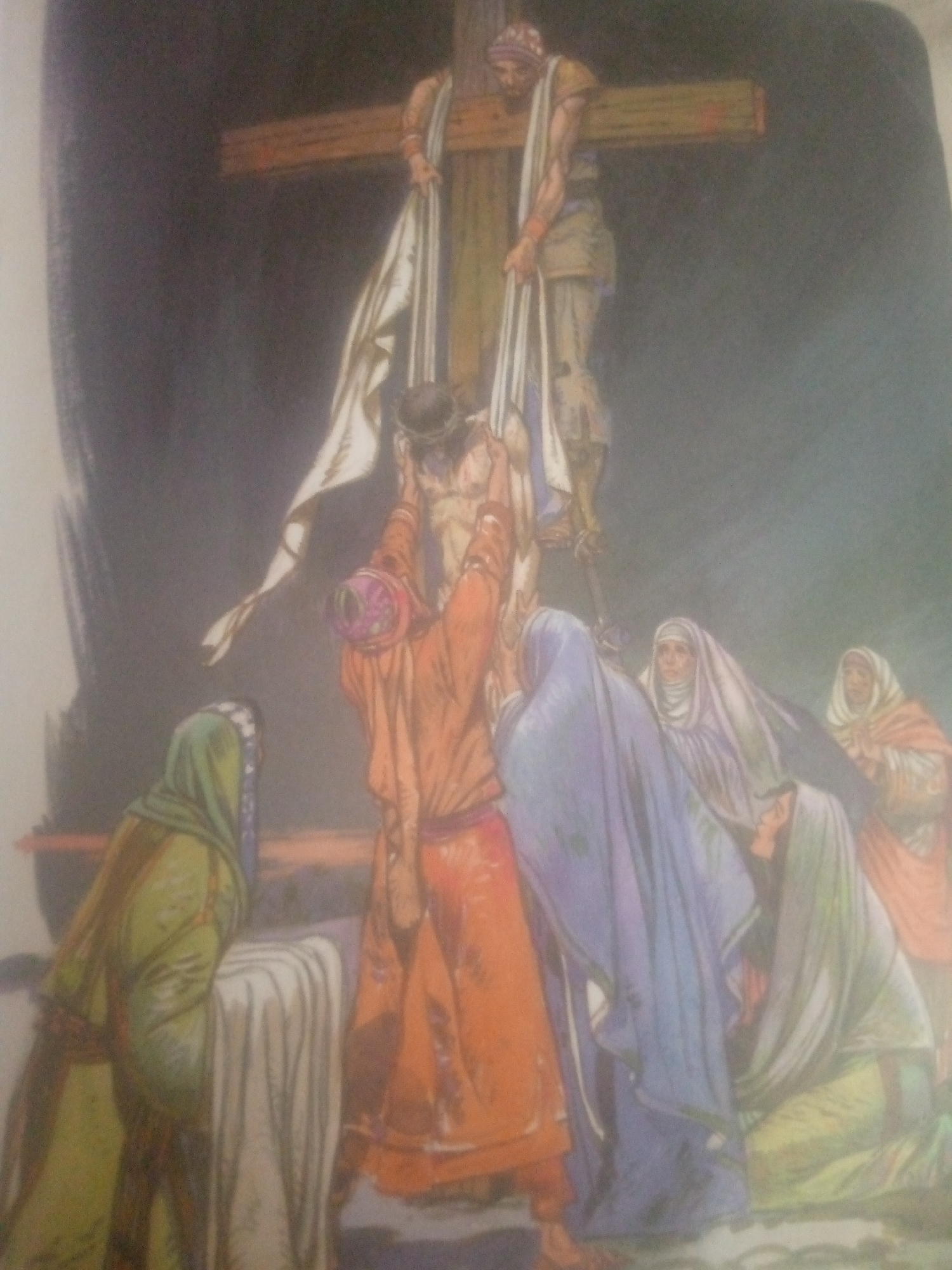 The image depicts a scene of the deposition from the cross. A man on the cross, appearing to be Jesus, is being lowered by two individuals. One man, positioned on the cross, is dressed in a light-colored garment and a headband. Another man, below him and to the left, is dressed in an orange robe and is actively involved in lowering the body. A group of women, dressed in various colored headscarves and robes, are gathered below, observing the event with solemn expressions. A woman in a green headscarf and robe is seated to the left, next to a white cloth. The background is dark and indistinct, suggesting an outdoor setting. There is no identifiable indication of the city of Jauro Yinu, Nigeria, in this image.