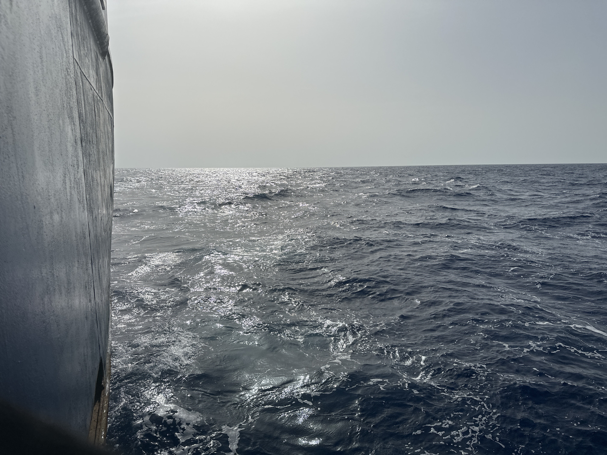 A view from a ship at sea. The port side of the ship is visible in the left foreground. Beyond the ship, the ocean stretches to the horizon under a bright, hazy sky. The water is dark with whitecaps and reflected sunlight. The location is unknown.
