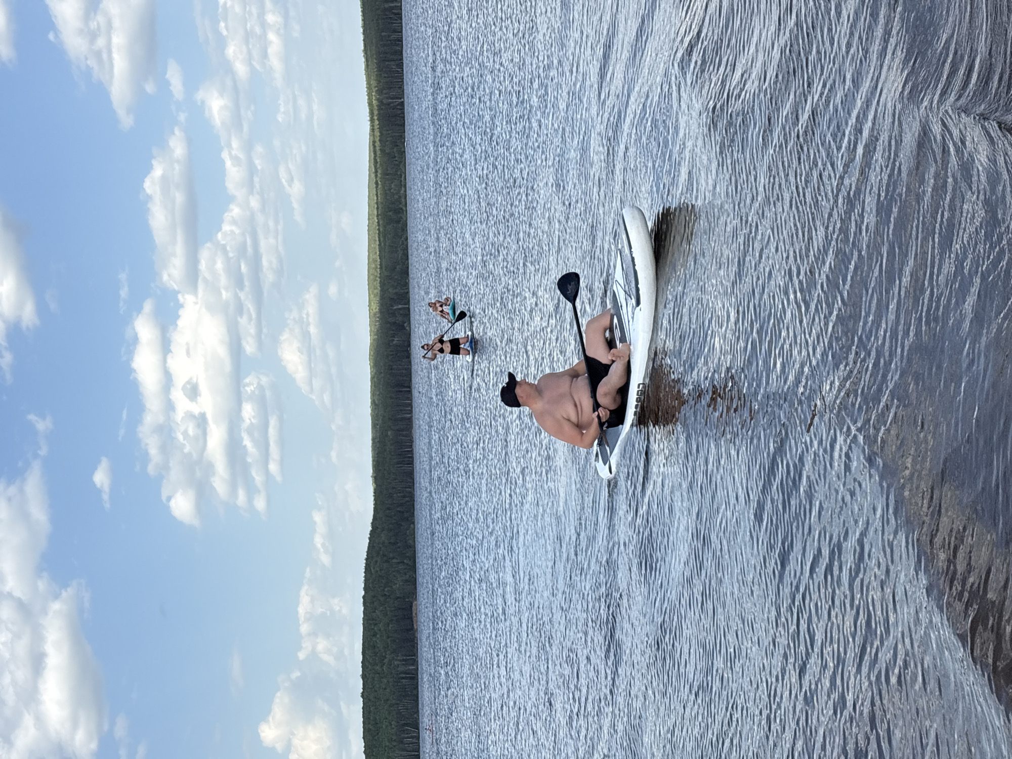 The image shows a man lying on a white paddleboard in a large body of water. He is wearing a black baseball cap and black shorts. A paddle lies next to him on the board. In the distance, two other people are standing on paddleboards. A forest lines the opposite shore. The sky is blue with scattered white clouds. The location is likely the city of Chaikovsky, Russia.