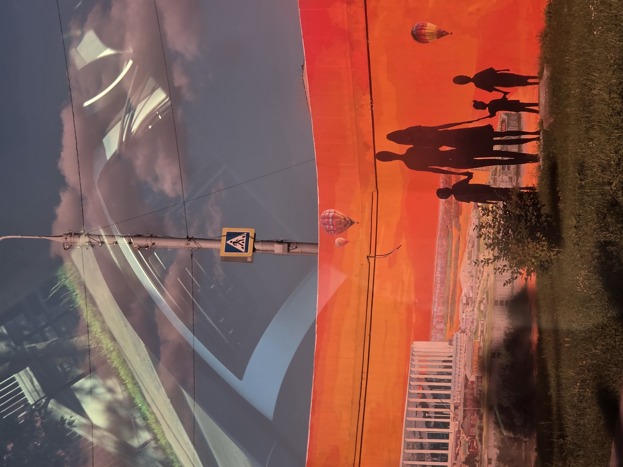 The image depicts a mural on a bright orange wall, with a silhouette of a family of four walking hand-in-hand in the foreground. Behind the family is a cityscape with a river and a modern white building. Hot air balloons are visible in the sky. Above the mural, the sky is visible with clouds and a lamppost with a pedestrian crossing sign. Reflections of what appear to be a highway or bridge structure are superimposed onto the sky. The scene is likely located in Nizhny Novgorod, Russia, given the distinctive architecture of the white building, which resembles the Nizhny Novgorod Stadium, and the presence of elements typical of Russian cityscapes. The overall impression is a vibrant and somewhat surreal urban scene, blending art with reality.