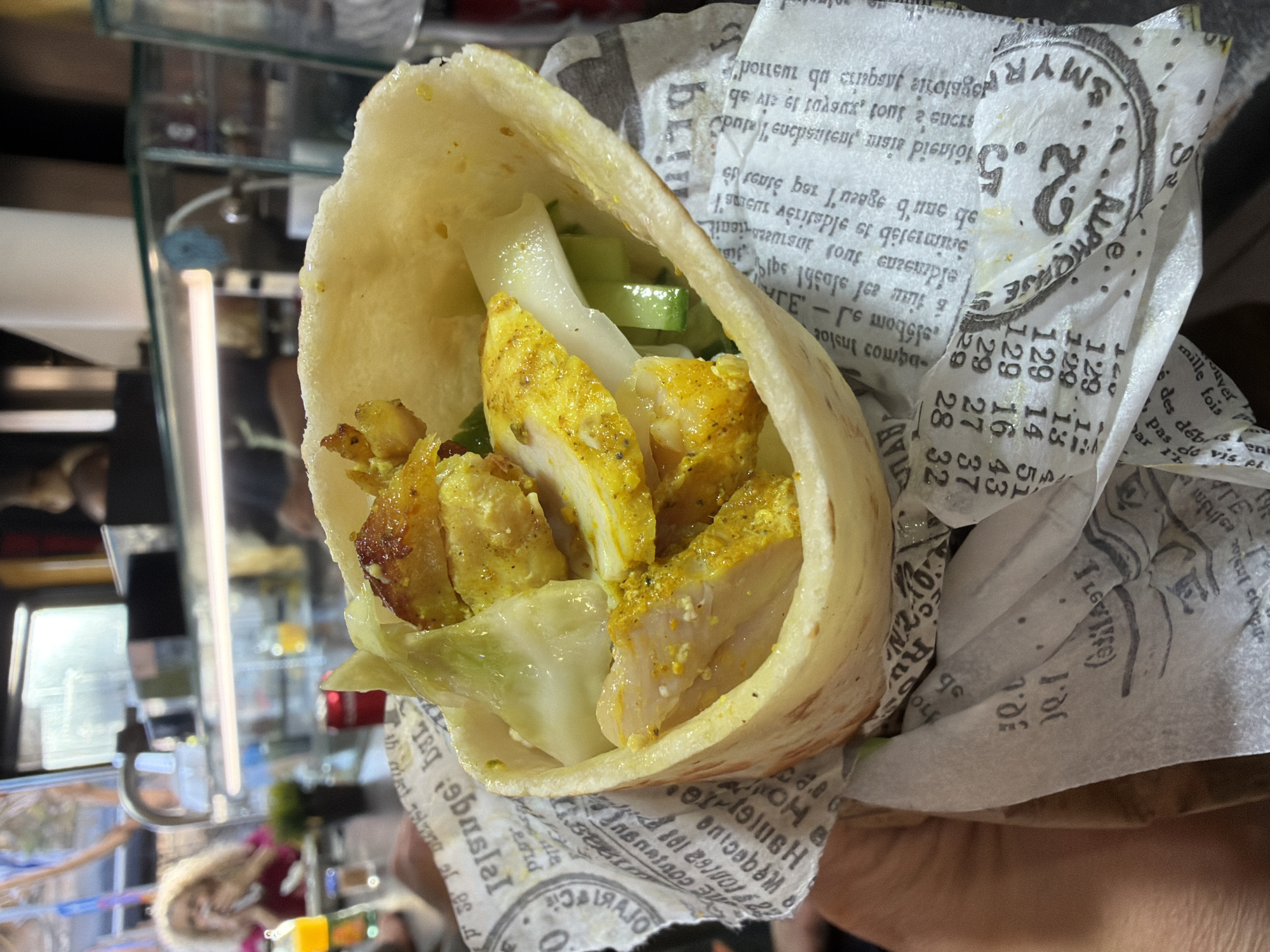 The image captures a close-up shot of a chicken shawarma wrap, likely from a street food vendor or restaurant in Tel Aviv, Israel. The wrap, filled with seasoned chicken, cucumber, and white sauce, is held in someone's hand. The wrap is encased in printed parchment paper. The background shows a blurred scene of a food establishment with glass-enclosed shelves.